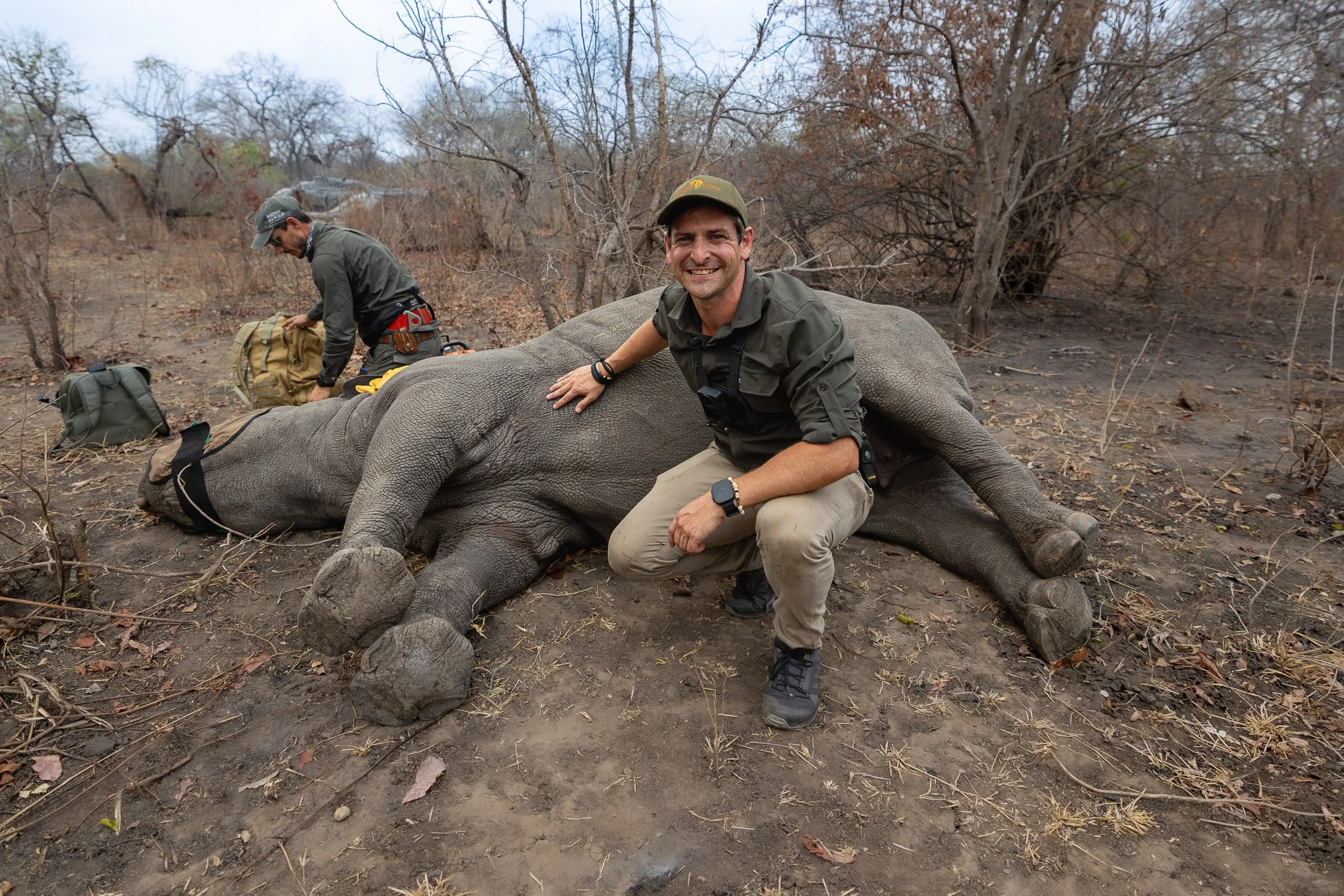 Marno Jooste beside a temporarily immobilised rhinoceros in Liwonde National Park, Malawi, with a wildlife veterinarian and helicopter in the background.