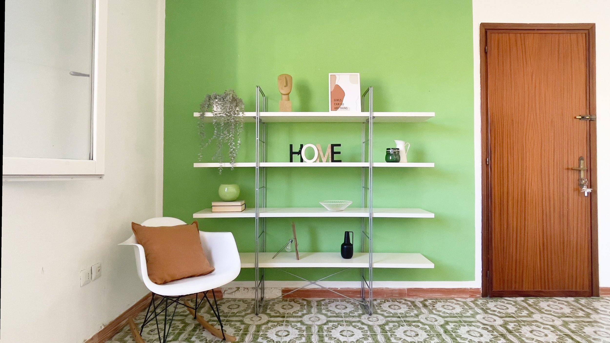 Soggiorno con sedia bianca e cuscino marrone, mensole bianche su fondo verde con decorazioni e oggetti ornamentali, porta di legno a destra.