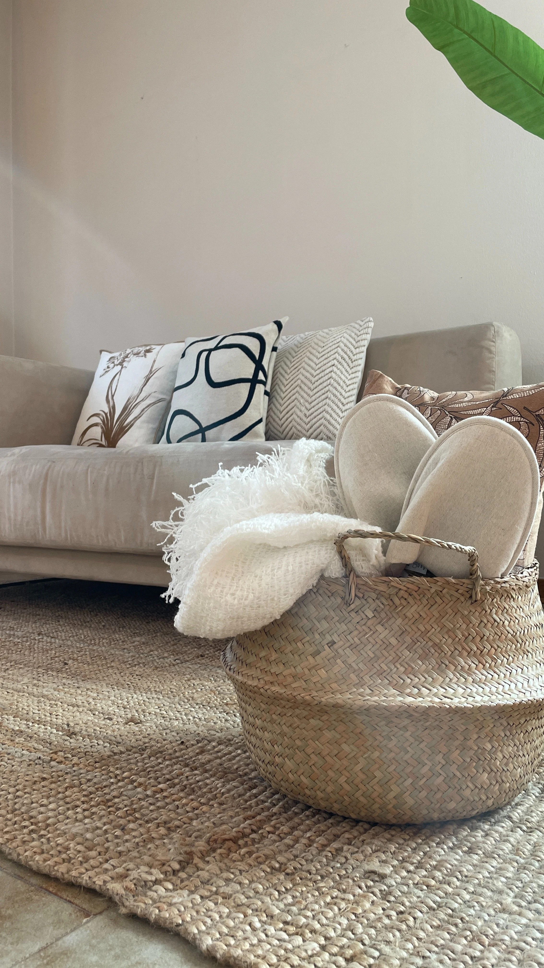 Soggiorno con divano beige, cuscini decorativi, cesta intrecciata contenente pantofole e coperta, pianta decorativa e tappeto di juta.