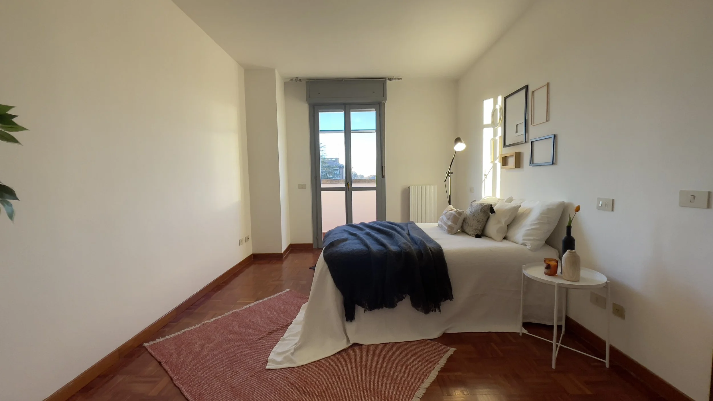 Camera da letto moderna con letto matrimoniale, coperta scura, tappeto arancione e tavolino con decorazioni. Finestra luminosa e parete con cornici. Pianta nella parte sinistra.