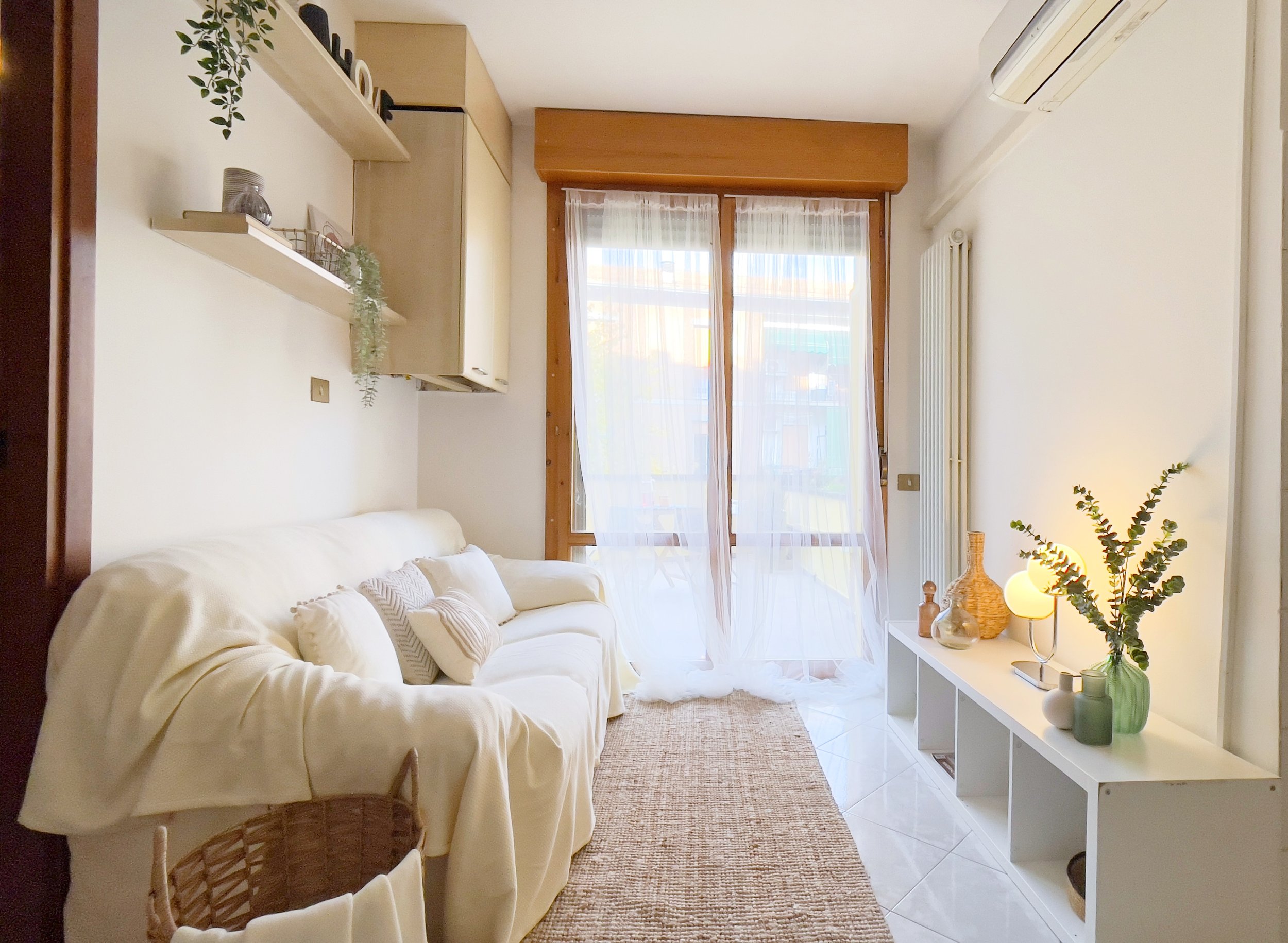 Soggiorno luminoso con divano bianco, tappeto in fibra naturale, scaffale con decorazioni e porta finestra con tende leggere.