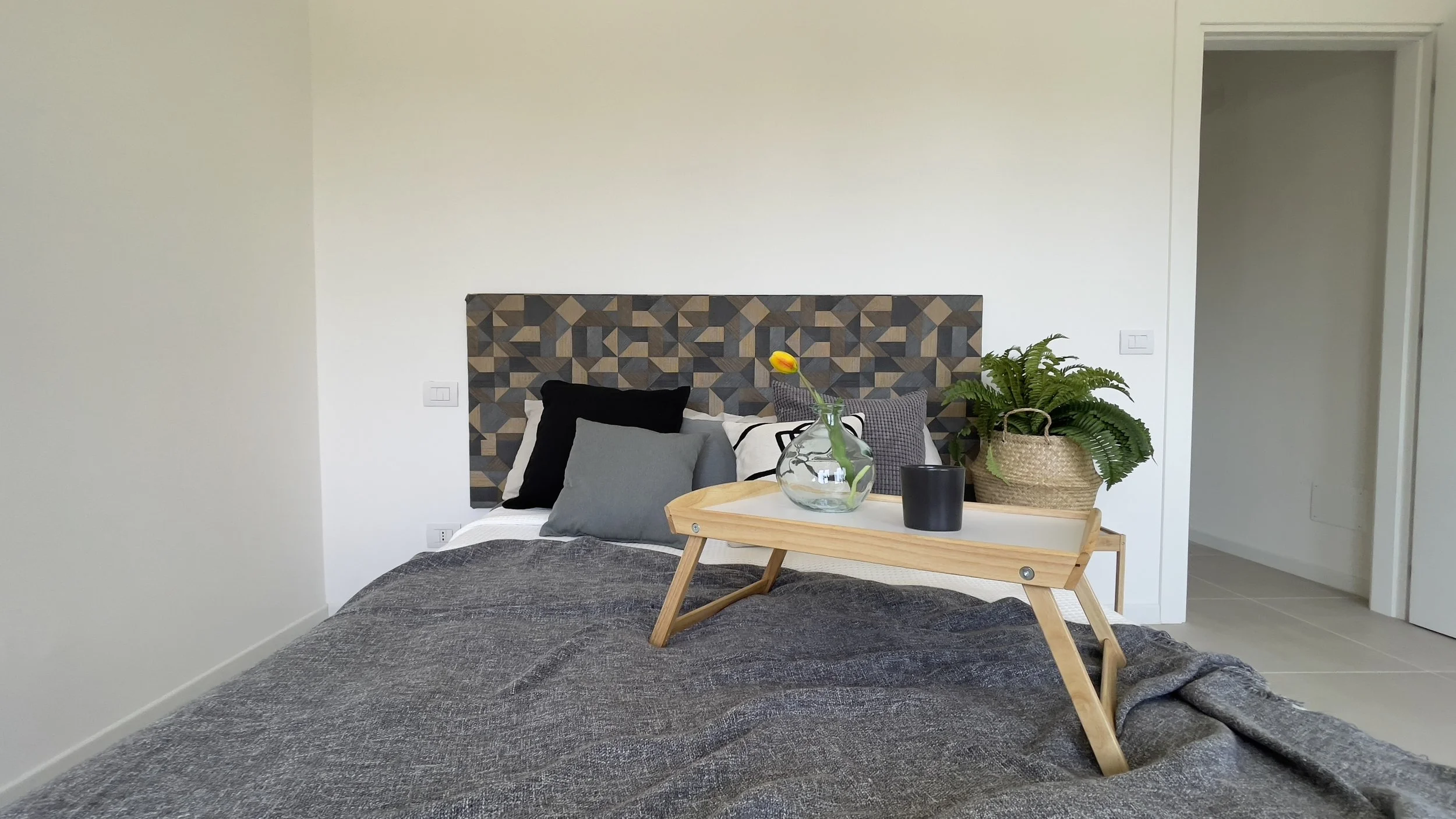 Camera da letto moderna con letto matrimoniale, testi a scacchi geometrici su testiera, cuscini e coperta grigi. Vassoio in legno con vaso di vetro e pianta in vaso accanto al letto.