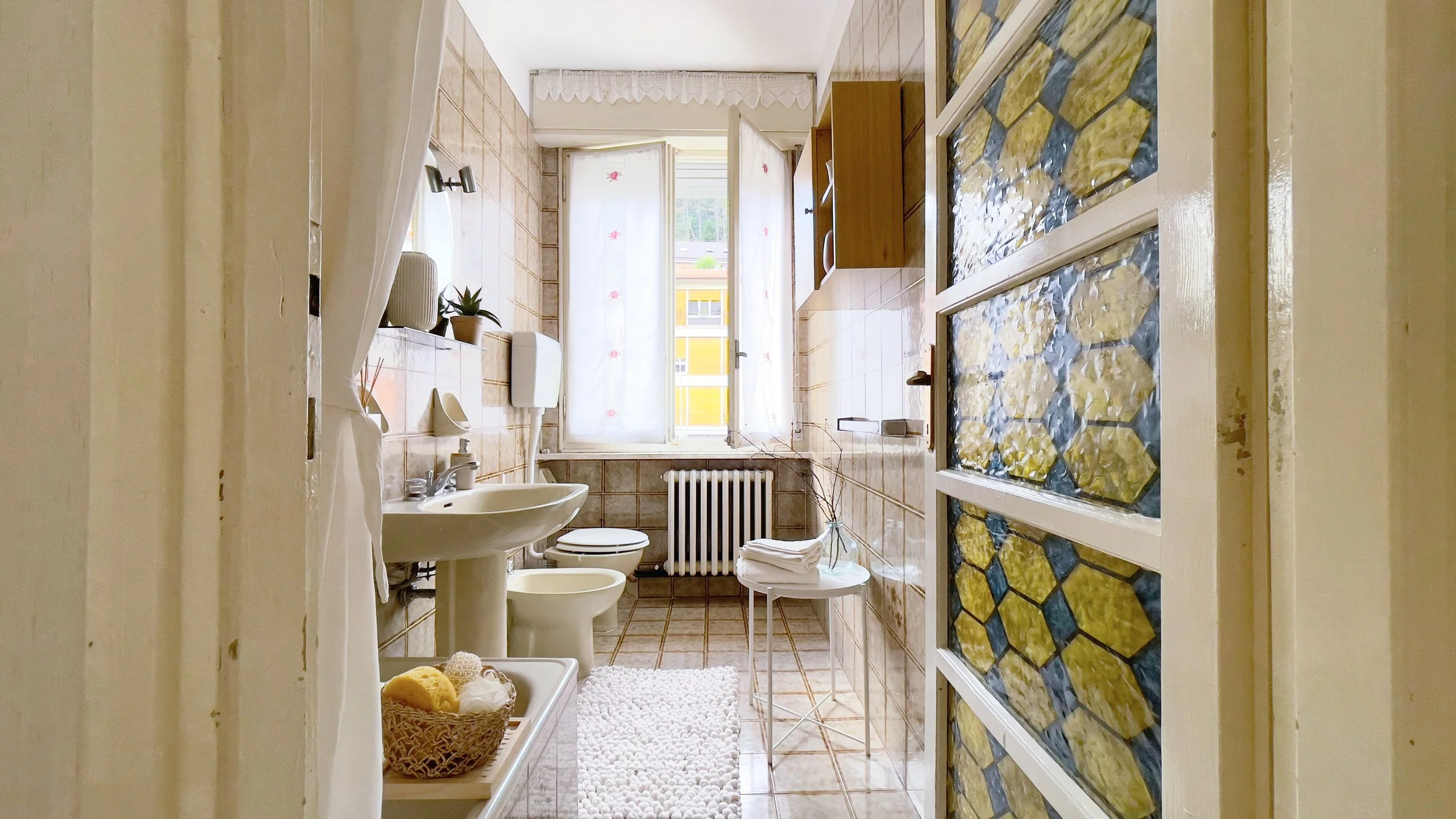 Bagno luminoso con lavandino, specchio, water, bidet, e finestra con tende. Cestino con spugne sul ripiano. Porta con vetri decorati.