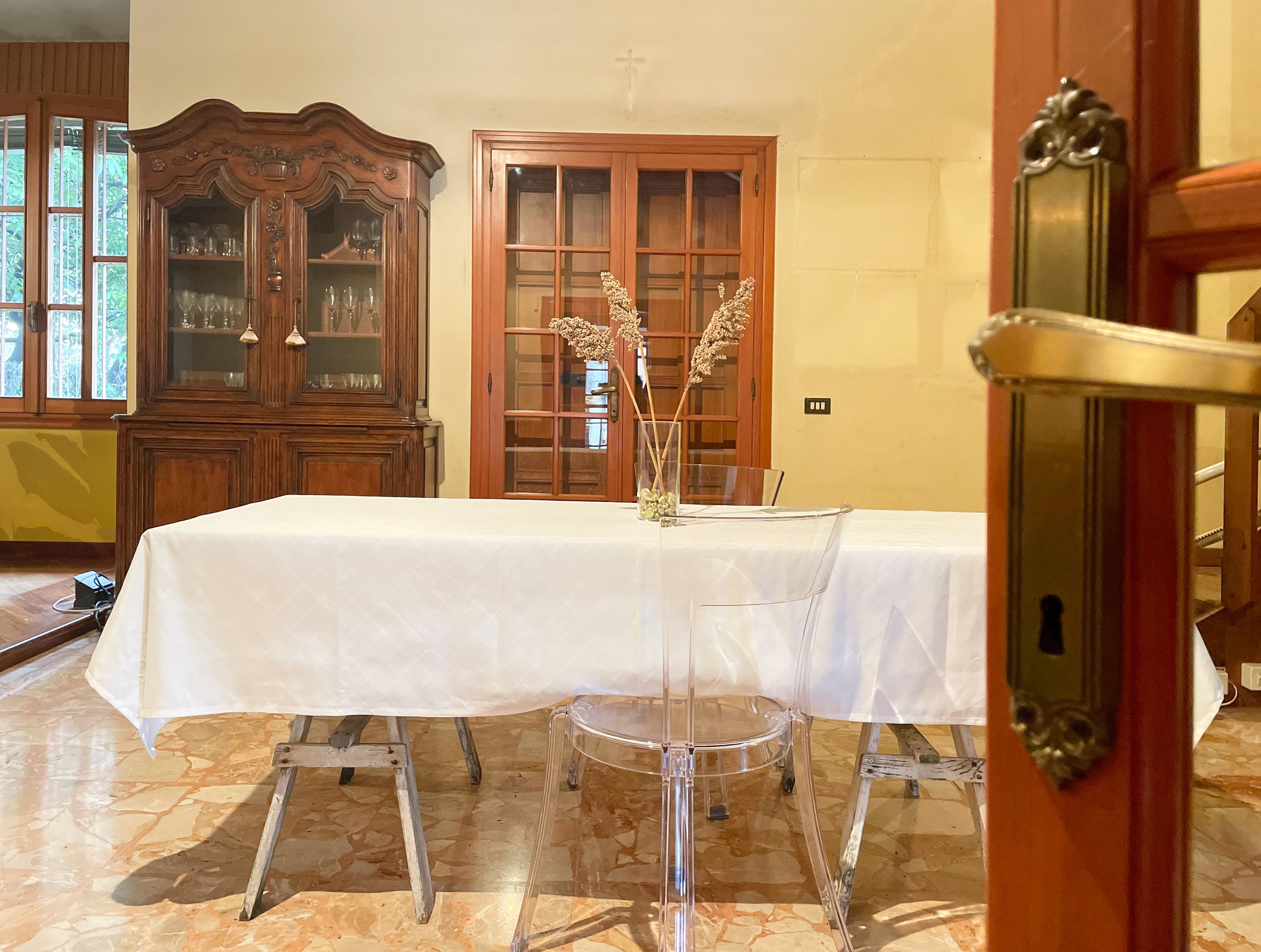 Interno di una stanza con un tavolo coperto da una tovaglia bianca, sedie trasparenti e un armadio antico sulla sinistra. Sul tavolo ci sono dei fiori secchi in un vaso. Sullo sfondo si vede una porta in vetro e legno.