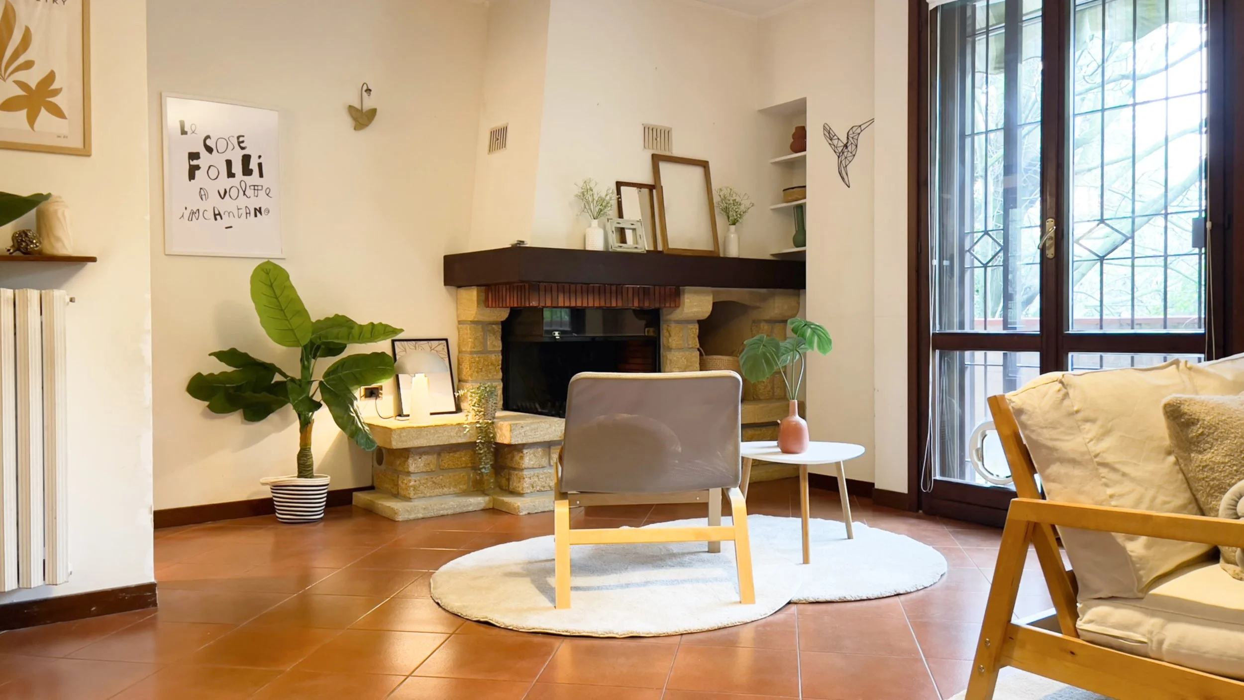 Interno di un soggiorno accogliente con camino in pietra, poltrona moderna, tavolino bianco, piante verdi e decorazioni minimaliste su pareti chiare. Grandi finestre lasciano entrare la luce naturale.