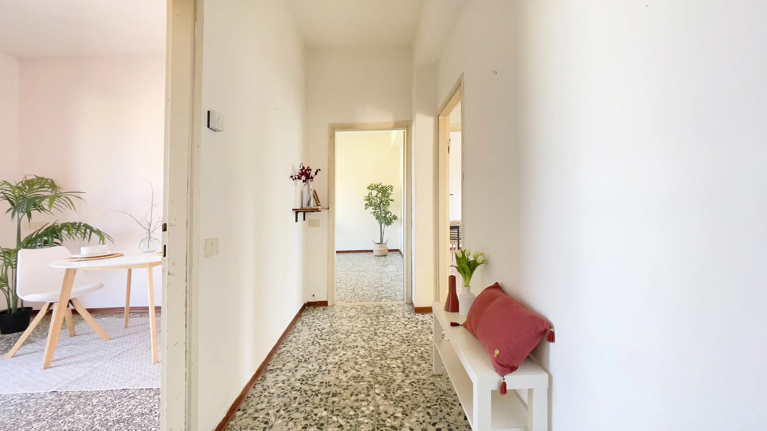 Corridoio luminoso con pavimento a mosaico, arredato con un tavolino e sedia a sinistra, piante in vaso, e una mensola con decorazioni. Vista di una stanza in fondo.