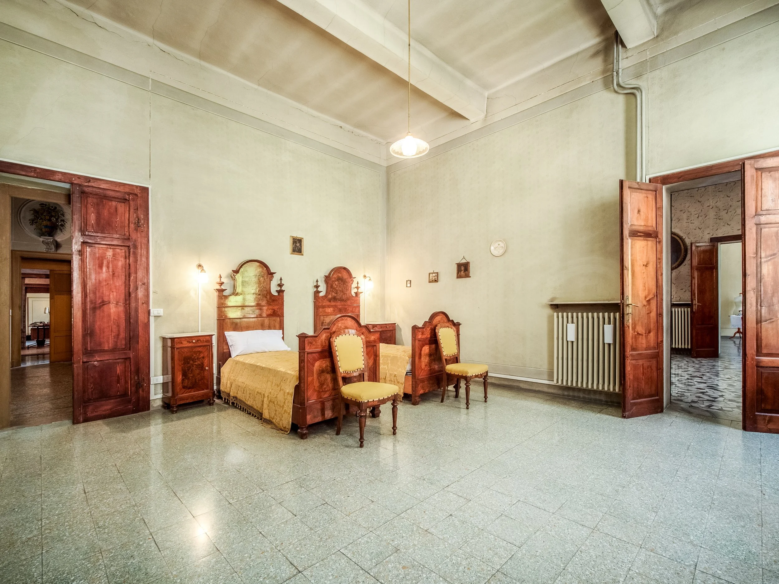 Camera da letto tradizionale con due letti singoli in legno, sedie abbinate, pareti color crema e pavimento piastrellato. Illuminazione soffusa e porte in legno.