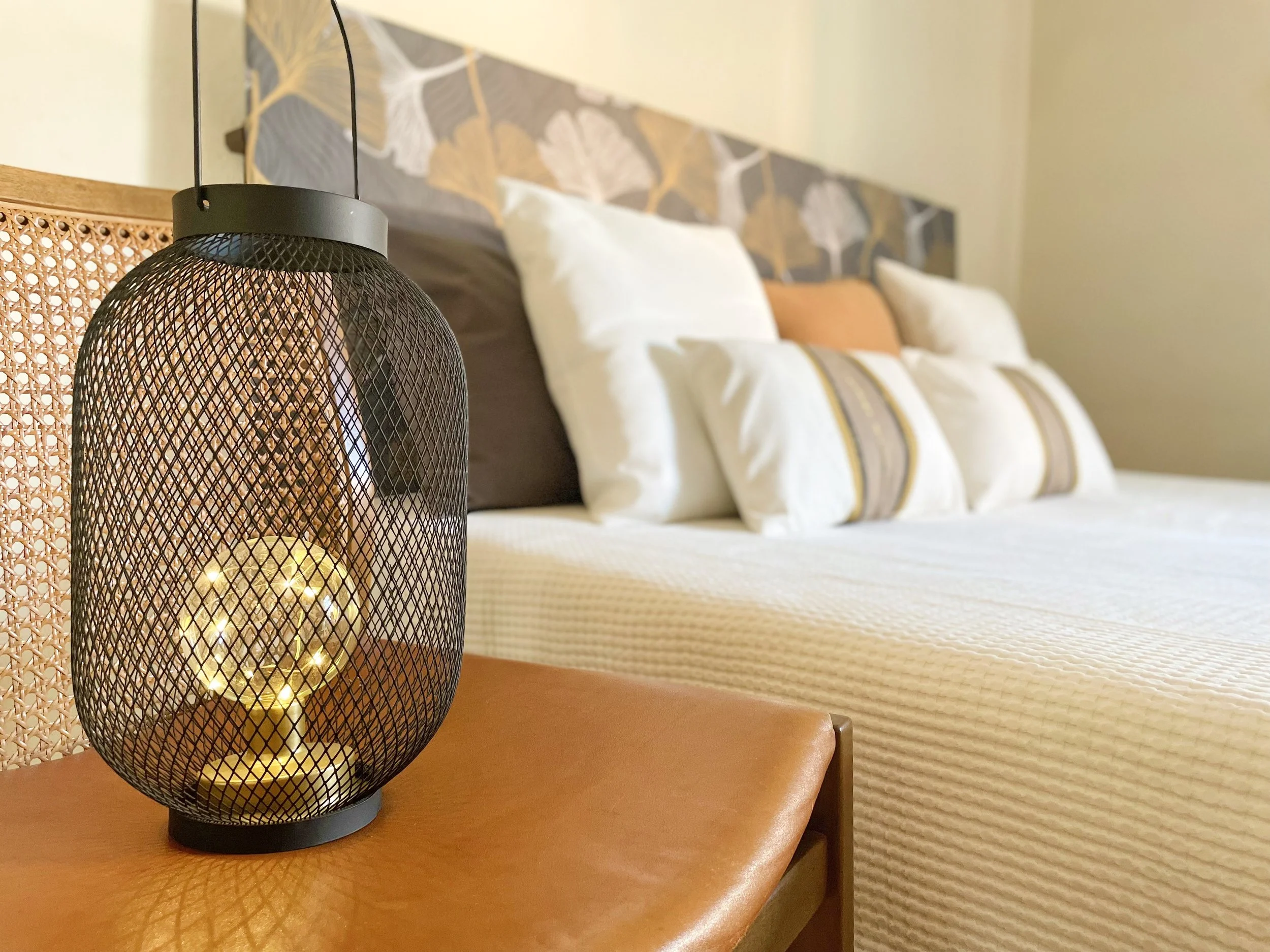 Lampada da tavolo a rete nera su un comodino con letto matrimoniale sullo sfondo, con cuscini bianchi e decorativi.