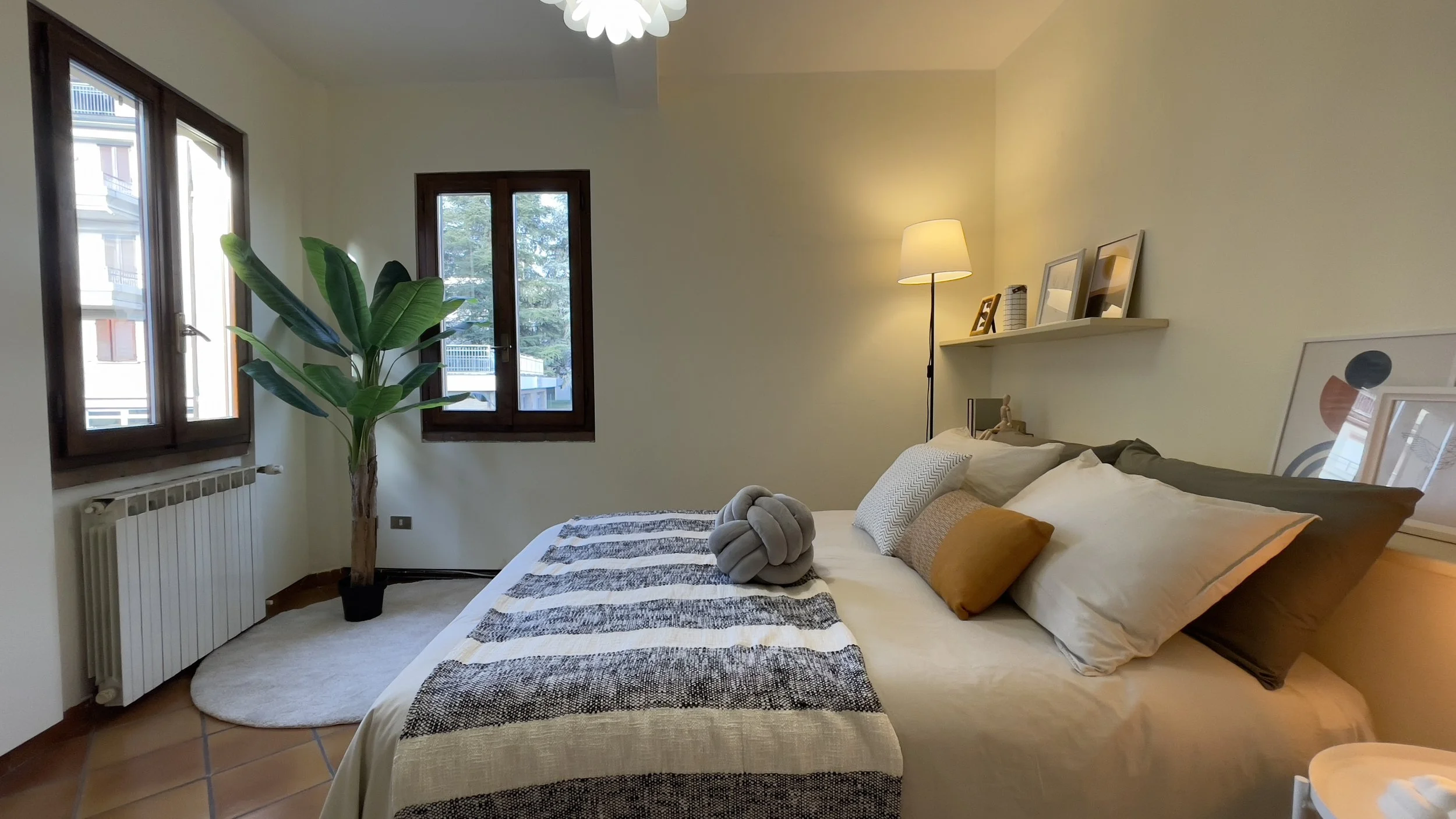 Camera da letto moderna con letto matrimoniale, cuscini variopinti, pianta decorativa, lampada da terra e quadri su mensola.