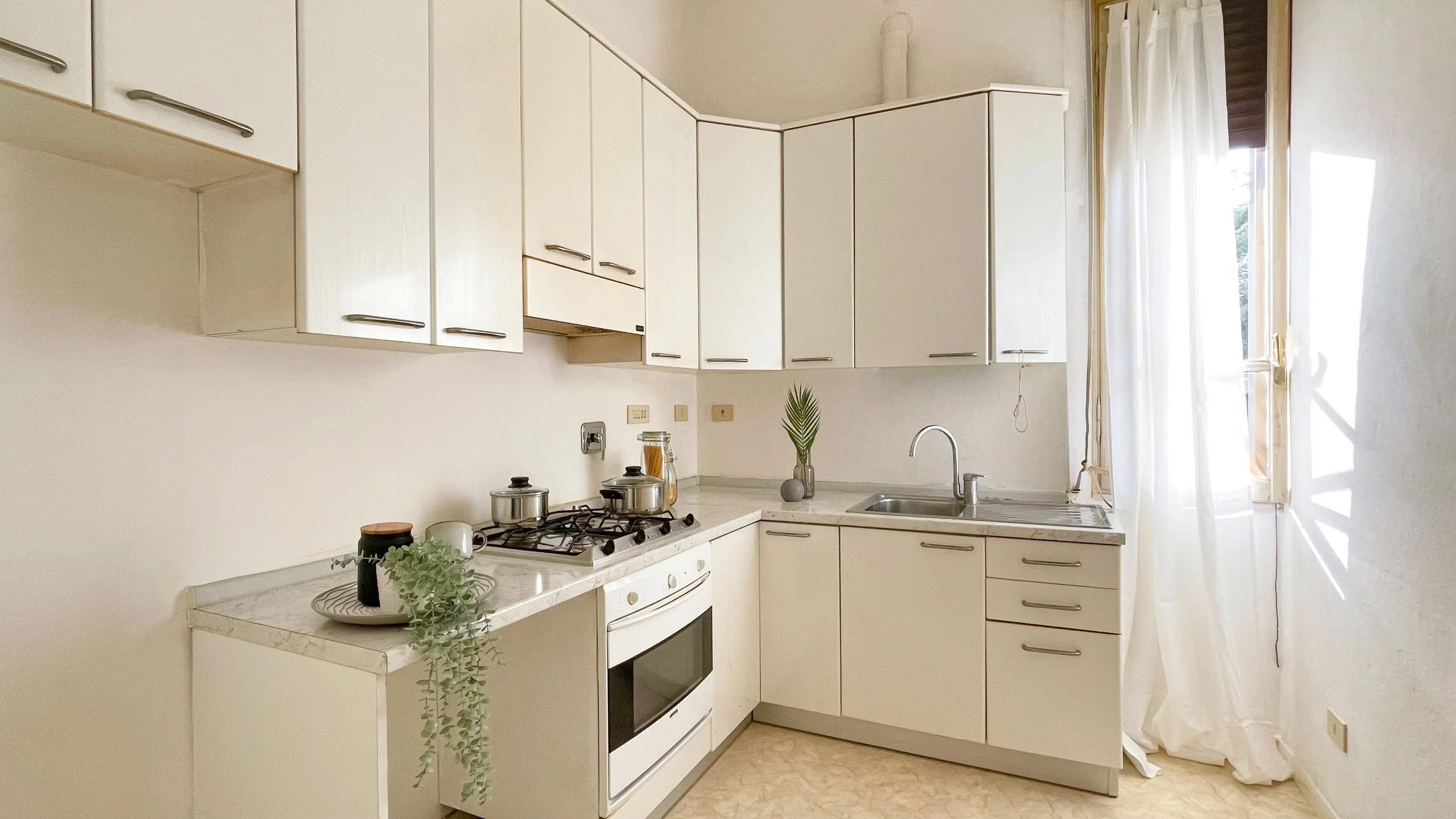Cucina moderna con mobili bianchi, pentole su fornelli a gas, lavello in acciaio inossidabile e finestra con tende bianche.