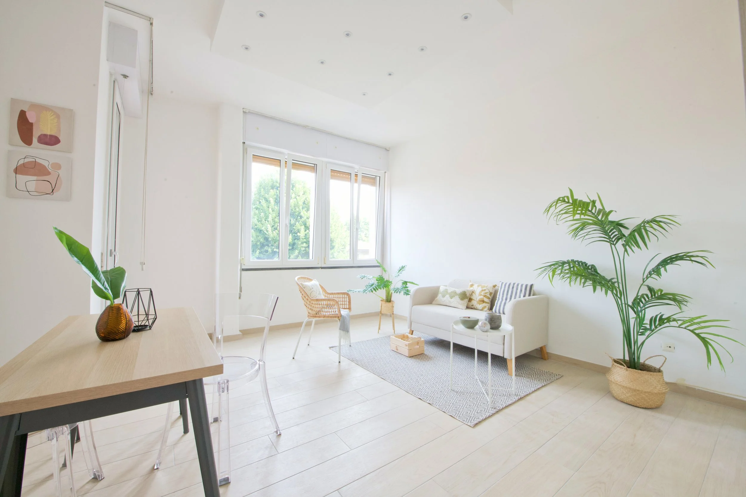 Stanza luminosa con arredamento minimalista, divano beige, piante verdi, tavolo e sedie trasparenti, decorazioni moderne sulle pareti, finestre ampie.