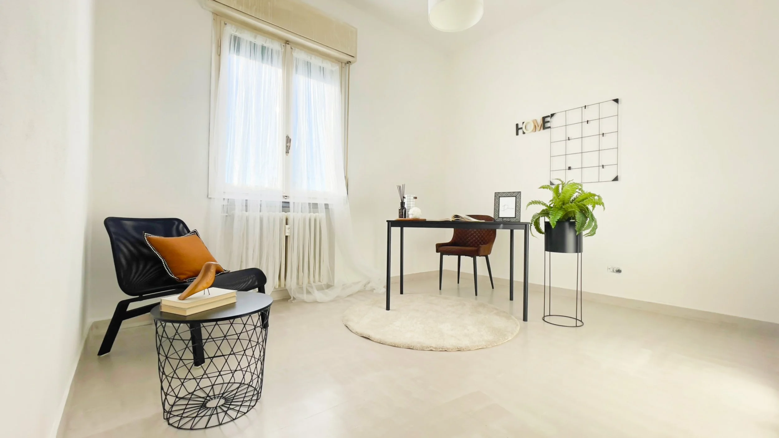 Interno di un ufficio moderno e minimalista con una sedia nera, tavolino con libri, scrittoio con oggetti decorativi e pianta in vaso, pavimento chiaro e finestra luminosa.