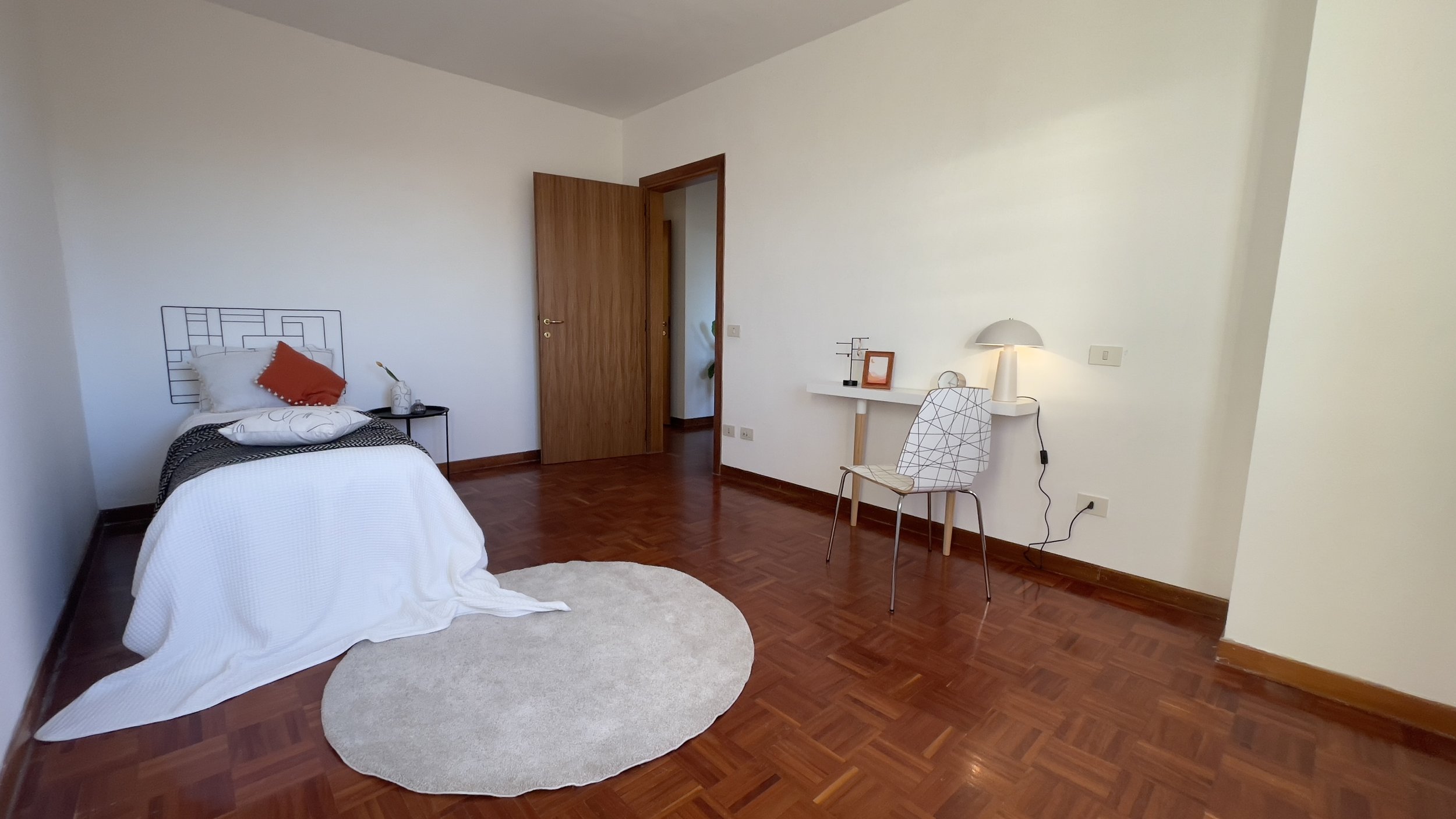 Camera da letto minimalista con letto singolo, coperta bianca, cuscini decorativi, tavolino laterale nero, pavimento in legno, tappeto rotondo chiaro, scrivania bianca con sedia moderna e lampada da tavolo.