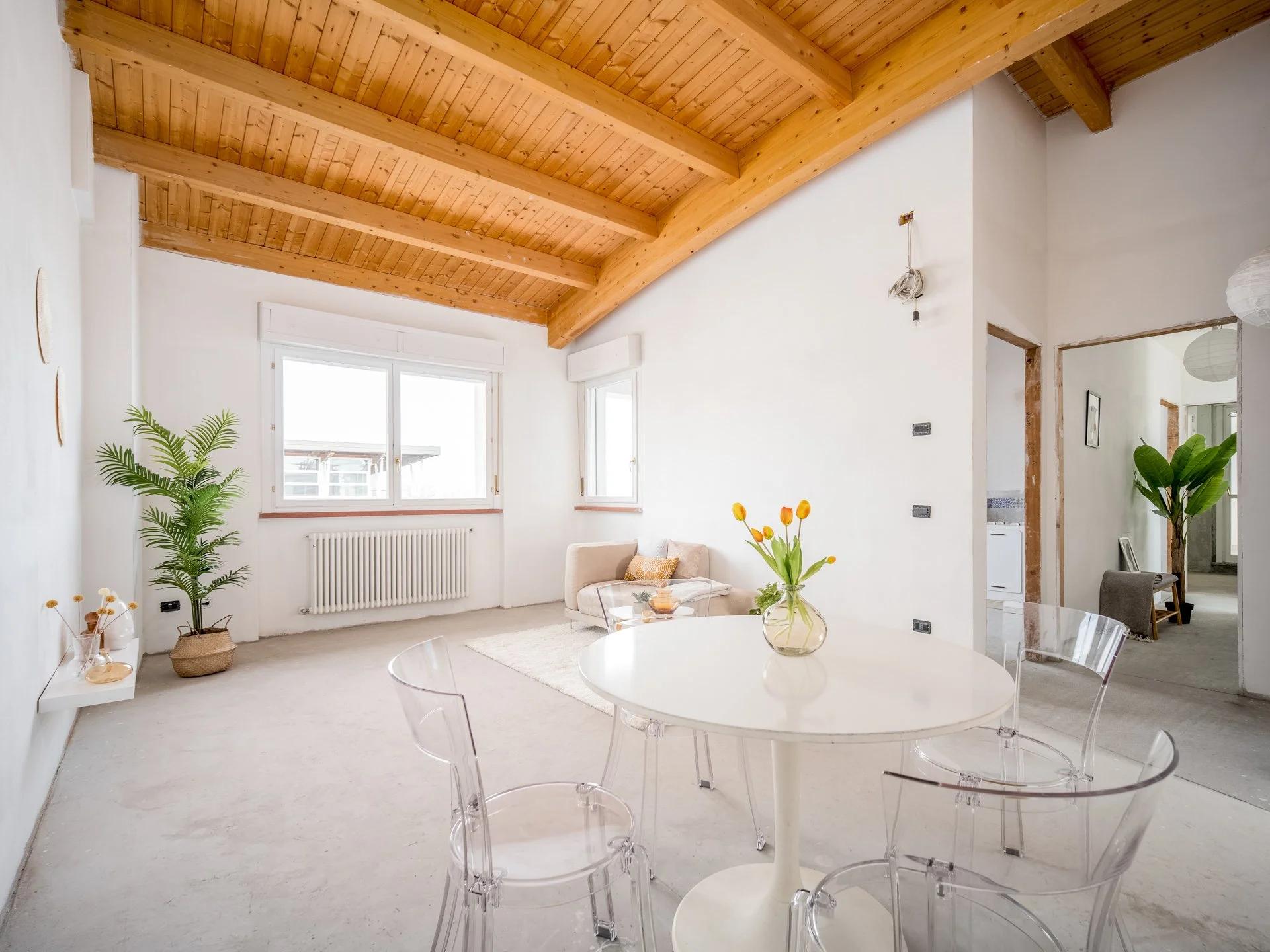 Interno di un soggiorno luminoso con tavolo rotondo bianco e sedie trasparenti, soffitto in legno, finestre ampie, pianta decorativa, e arredamento minimalista.