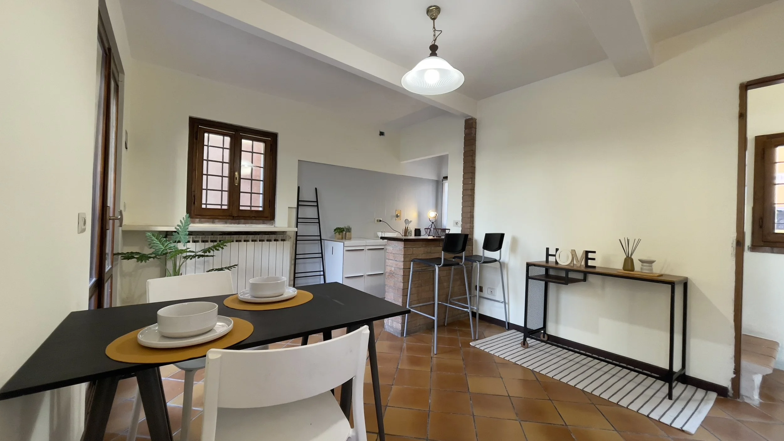 Interno di un soggiorno moderno con pavimento in cotto. Tavolo con sedie e stoviglie, cucina a vista con bancone in mattoni, e sgabelli. Decorazioni con piante e scritta 'HOME'. Finestre con cornici in legno.