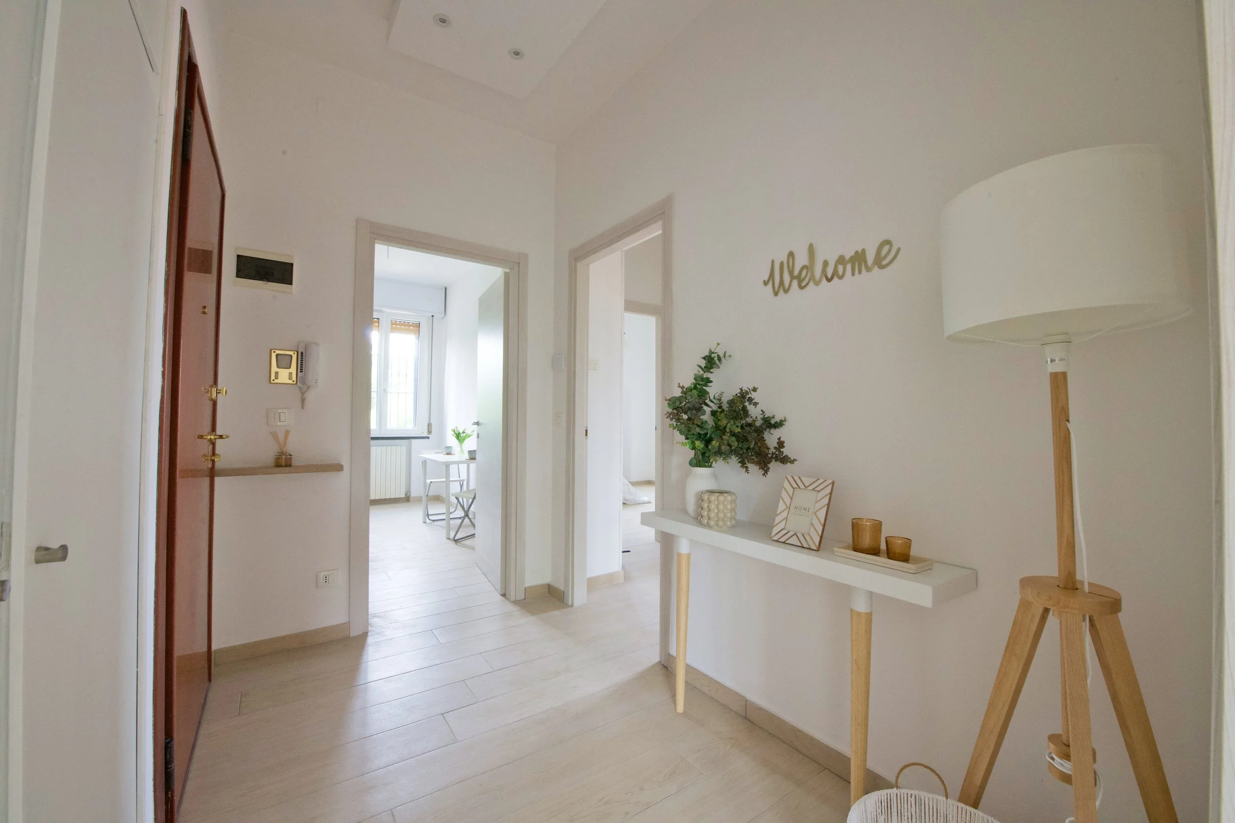 Ingresso di un appartamento con pareti bianche, tavolino con decorazioni e pianta, lampada da terra in legno e scritta "Welcome" sulla parete. Porta aperta conduce a sala illuminata.