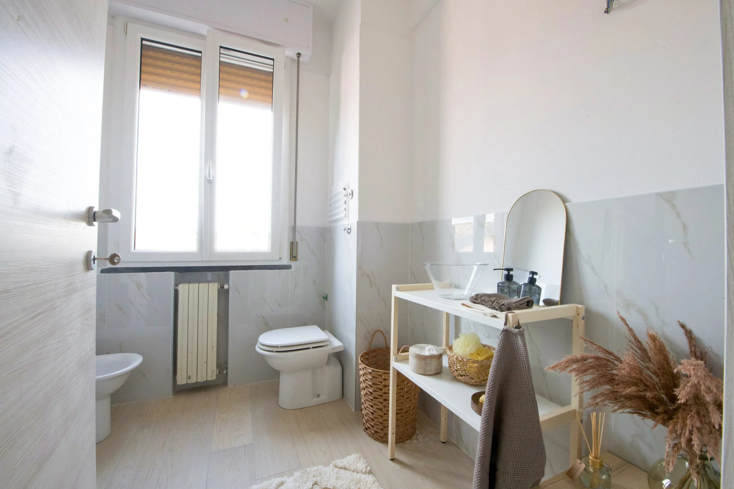 Bagno luminoso con finestra aperta, WC, bidet, scaffale bianco con oggetti da toeletta, specchio piccolo, piante decorative e cesto intrecciato.