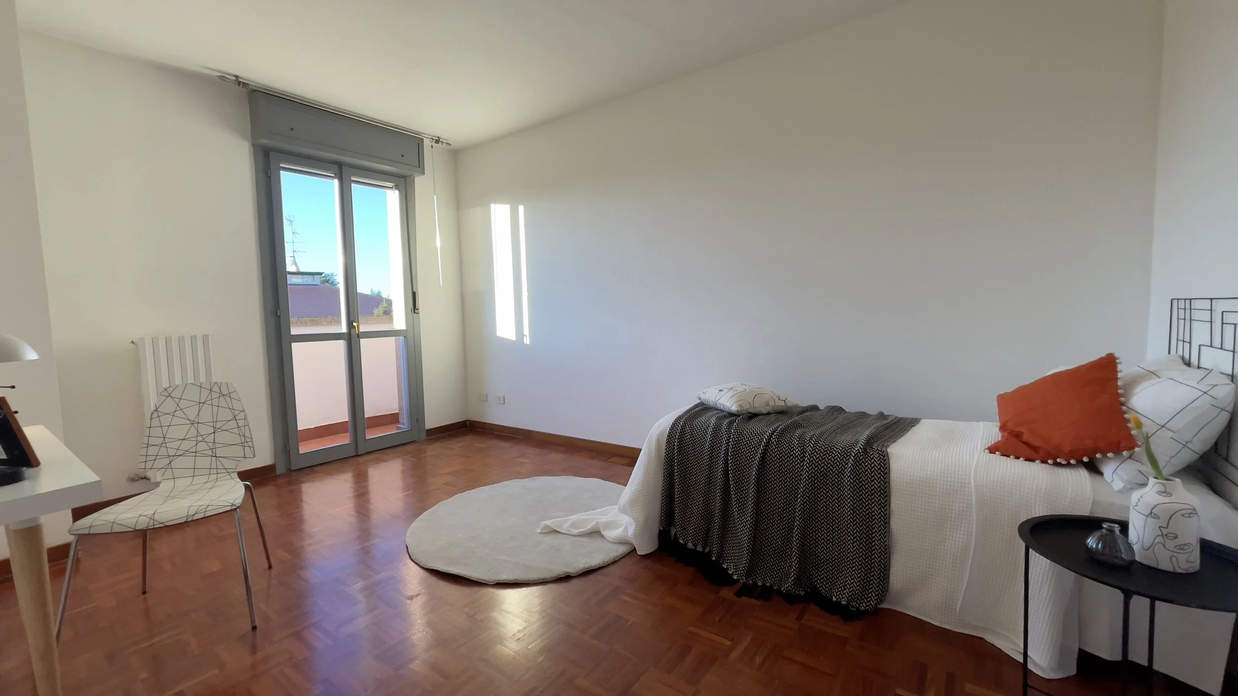 Camera da letto minimalista con parquet, letto singolo, cuscini colorati, tavolino nero, sedia, lampada e porta-finestra che dà su un balcone con vista esterna.