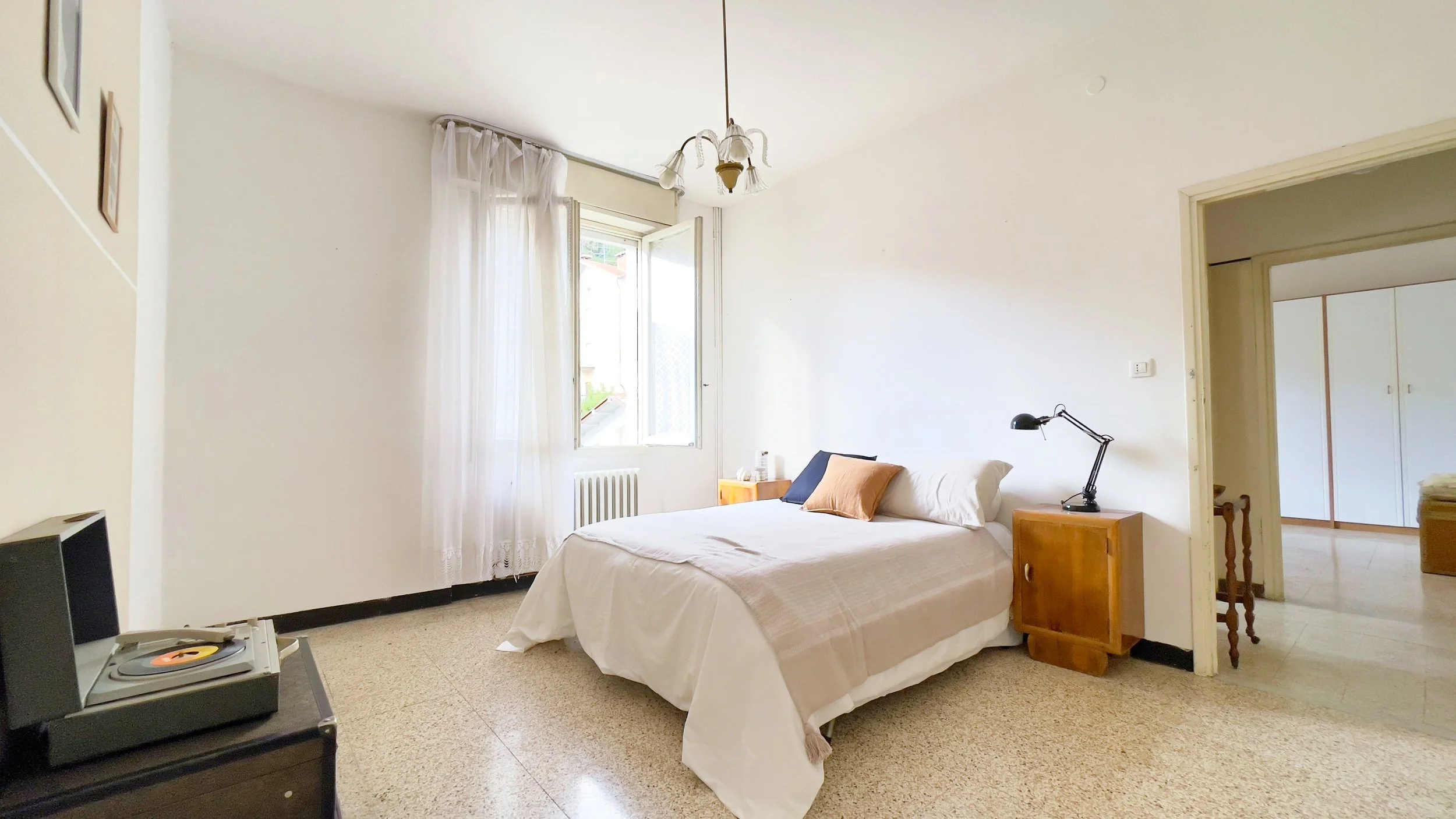 Camera da letto luminosa con letto matrimoniale, comodino in legno, lampada da tavolo nera e giradischi su mobile. Finestra con tende trasparenti e arredamento semplice.