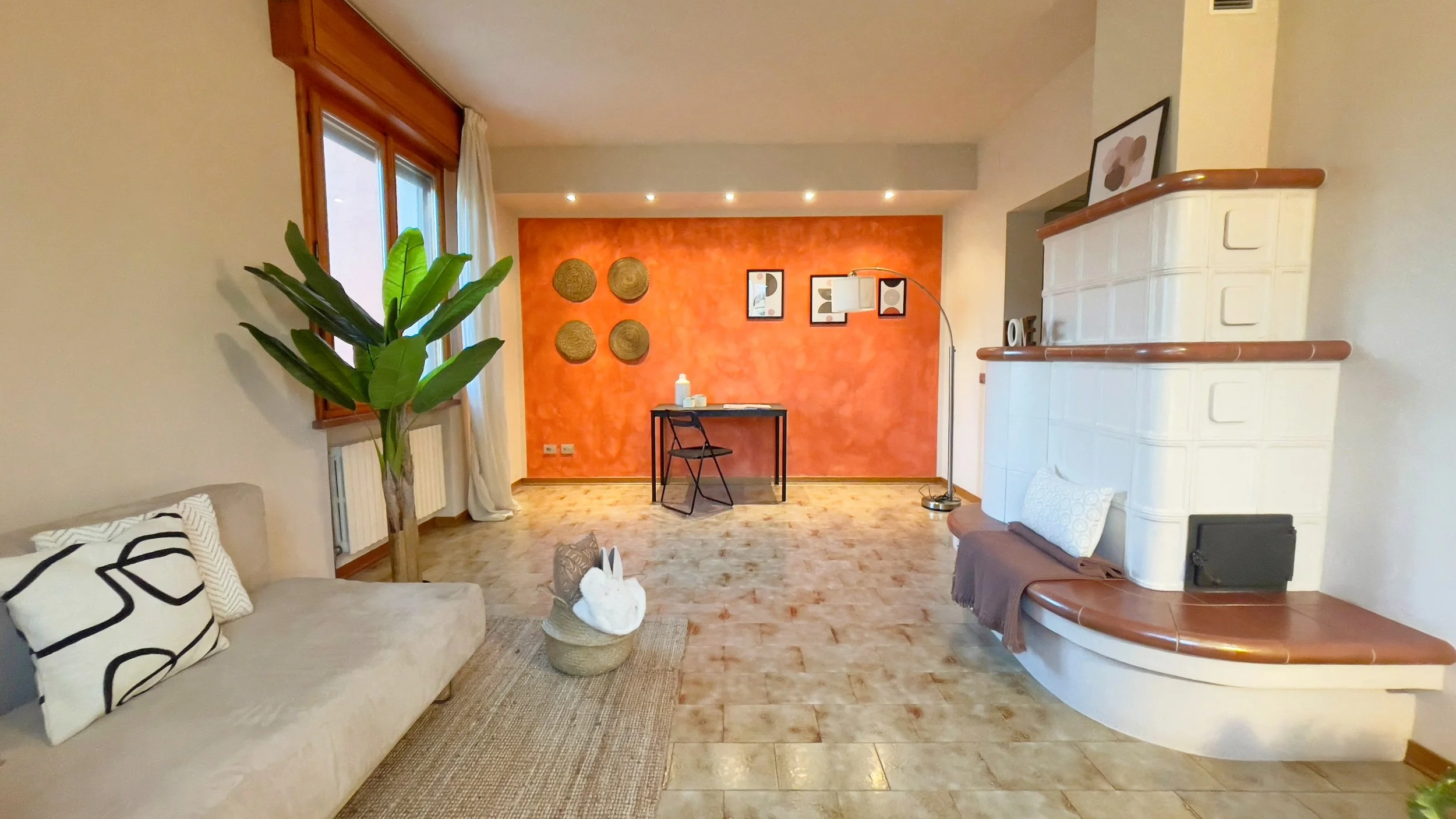 Interno di un soggiorno con parete arancione, tavolo nero e sedia pieghevole. A sinistra, divano con cuscini e pianta alta vicino alla finestra. A destra, stufa di design con panca curva e decorazioni sulle pareti.