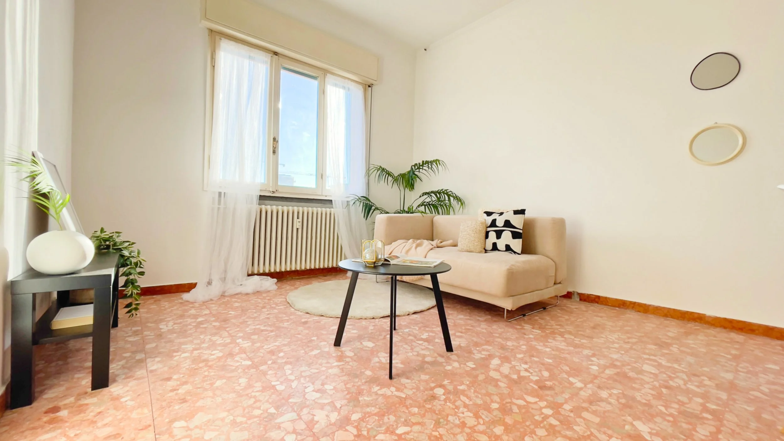 Soggiorno luminoso con divano beige, tavolino nero, cuscini decorativi e pianta verde. Pavimento in marmo e finestre con tende bianche. Specchi decorativi sulla parete bianca.