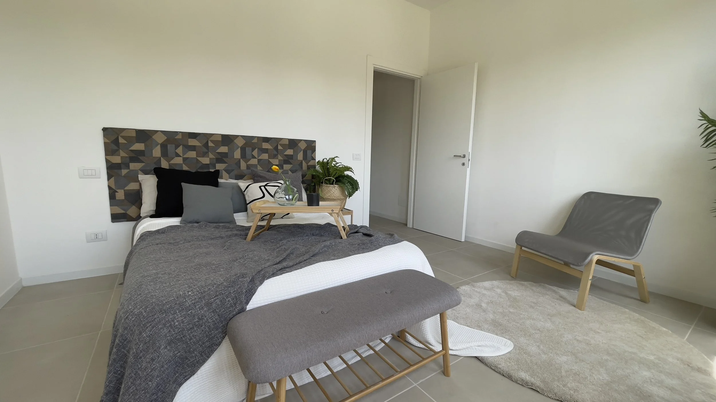 Camera da letto moderna con letto e coperta grigia, testiera geometrica, sedia grigia, pianta verde e tappeto chiaro.