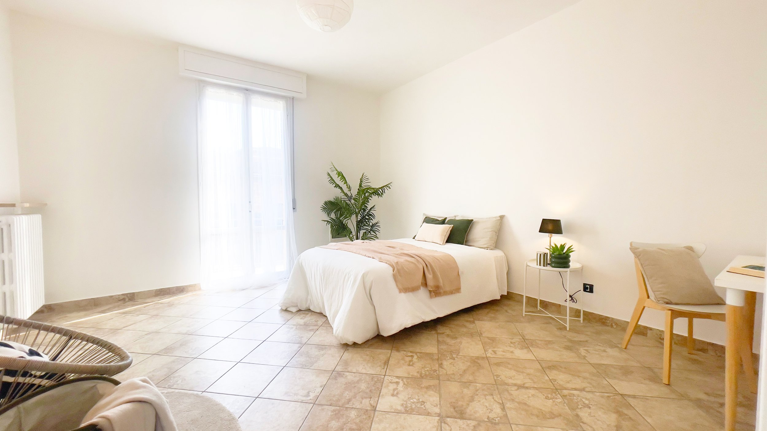 Camera da letto luminosa con letto matrimoniale, copriletto beige, piante in vaso, lampada da tavolo su comodino, sedia con cuscino e pavimento in piastrelle.