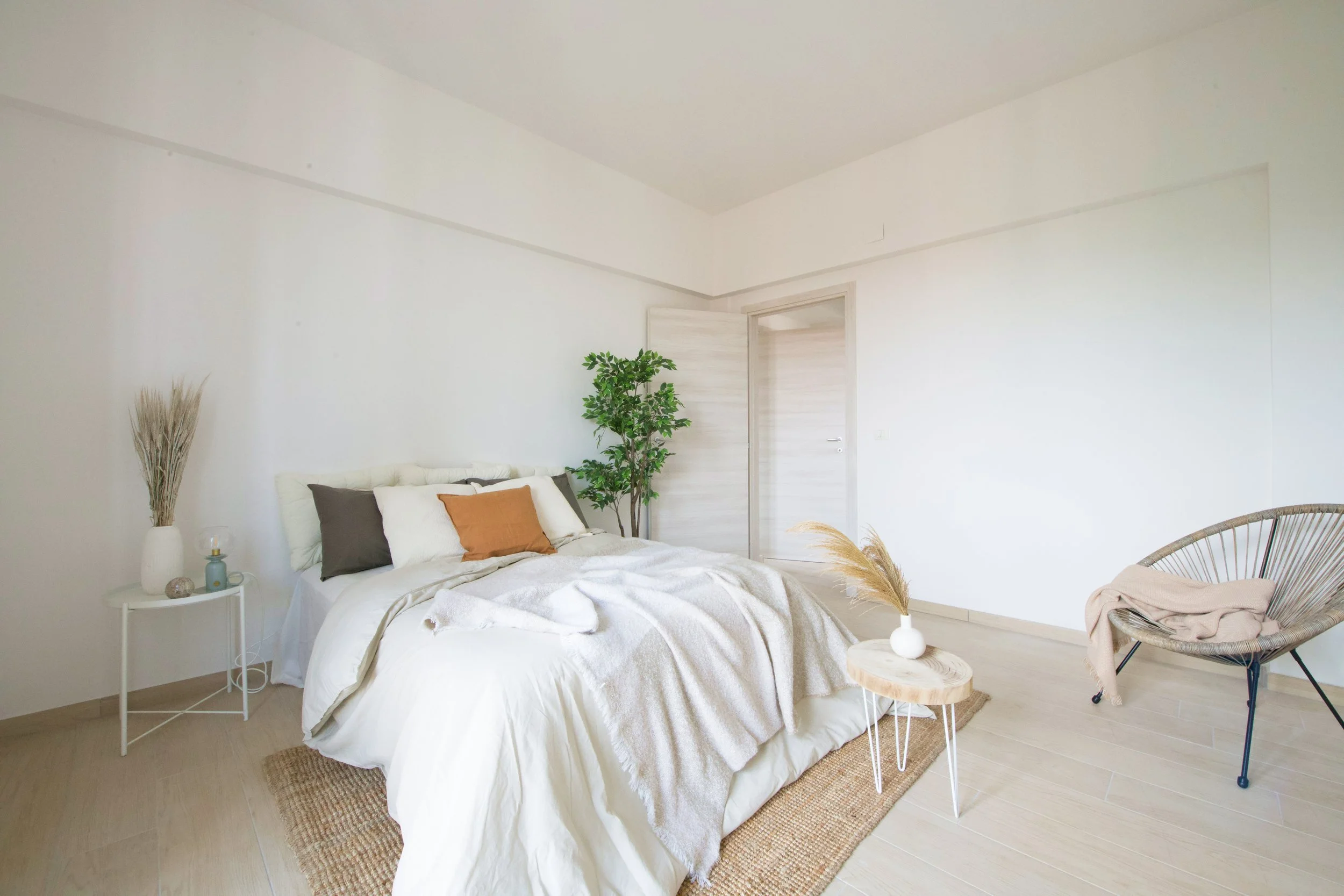 Camera da letto minimalista con letto, cuscini colorati, pianta verde, sedia e tavolino con decorazioni naturali.