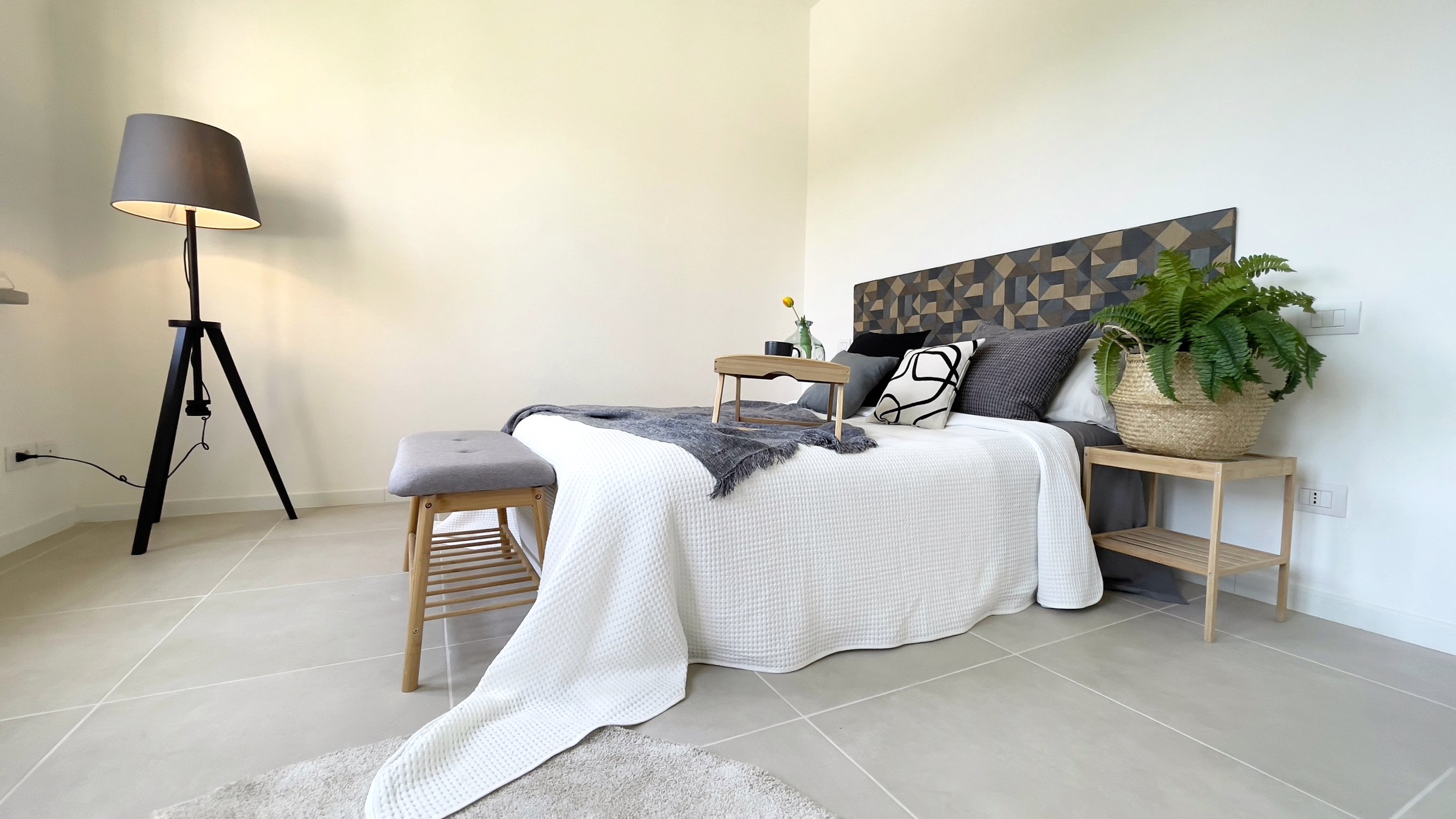 Camera da letto moderna con letto matrimoniale, lampada da terra, panca, coperta bianca, cuscini decorativi, tavolino con pianta e vassoio.