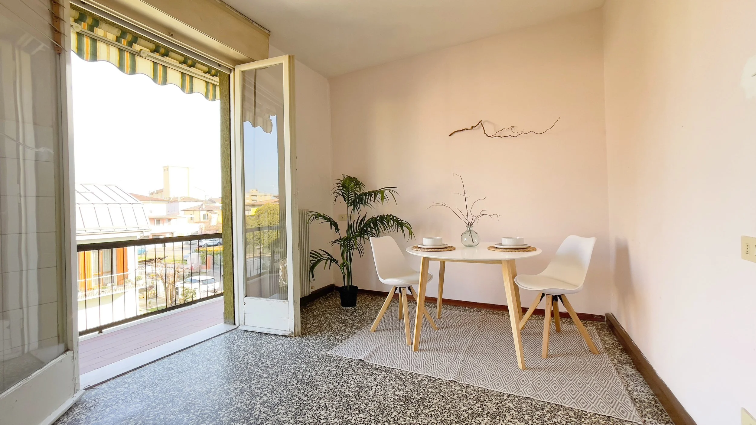 Interno di una stanza luminosa con un tavolo rotondo apparecchiato per due persone, due sedie bianche, una pianta verde in un vaso e una porta aperta su un balcone.