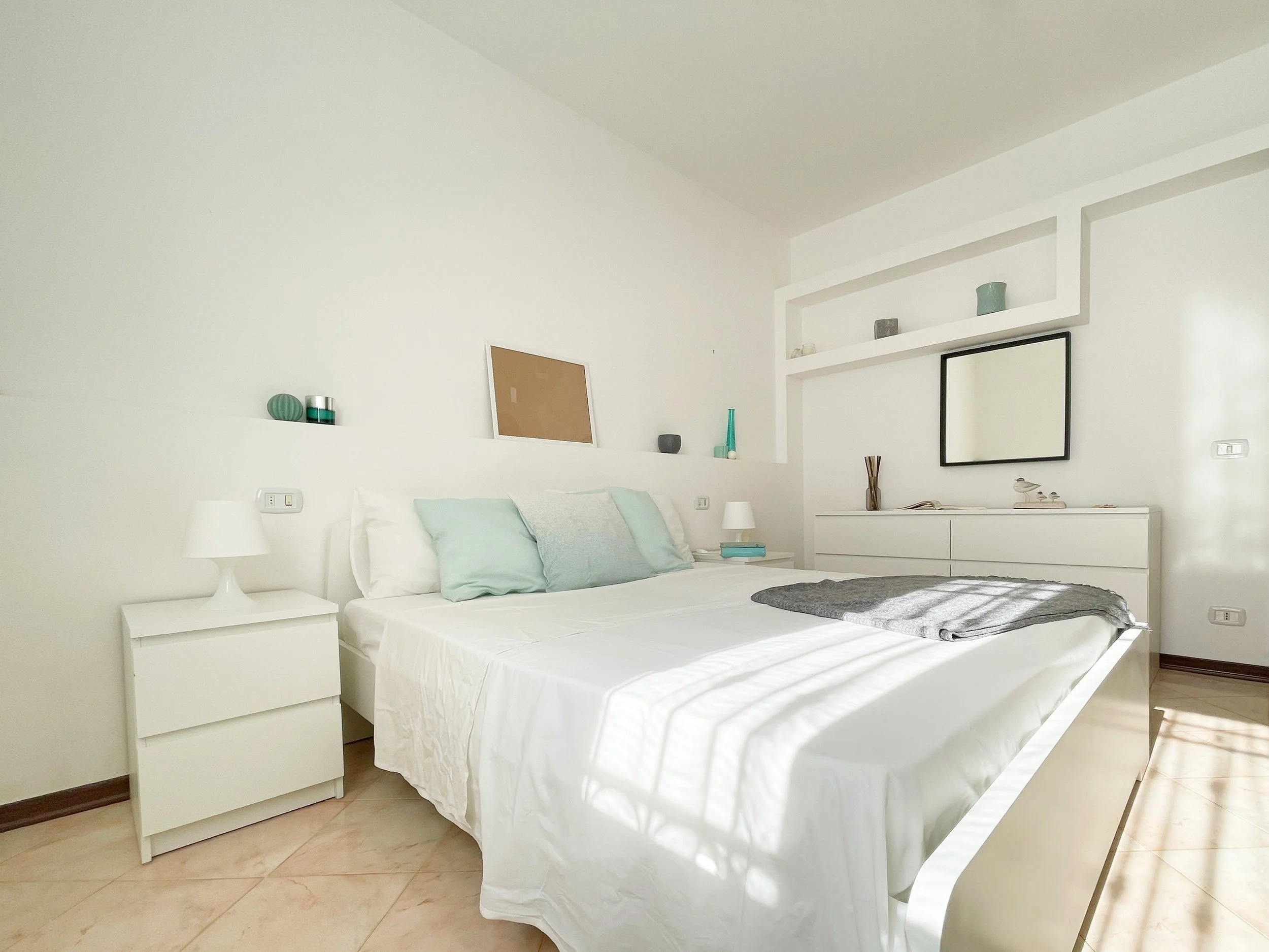 Camera da letto moderna con letto matrimoniale bianco, cuscini in tonalità pastello, comodino bianco con lampada, cassettiera, scaffali integrati e decorazioni minimaliste.