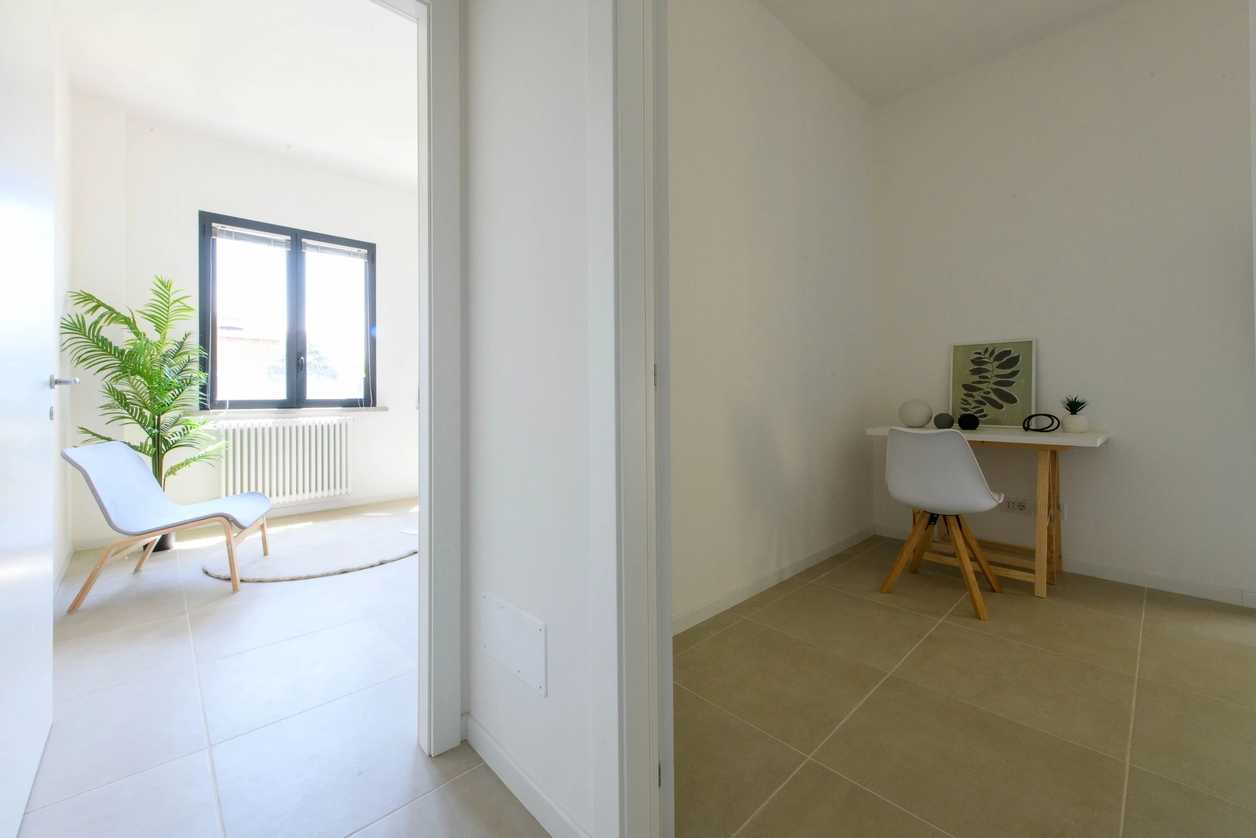 Interno di una casa moderna con pareti bianche, due stanze visibili. Una ha una sedia bianca e una grande pianta vicino a una finestra con telaio nero. L'altra ha una scrivania bianca, una sedia moderna e decorazioni minimaliste.