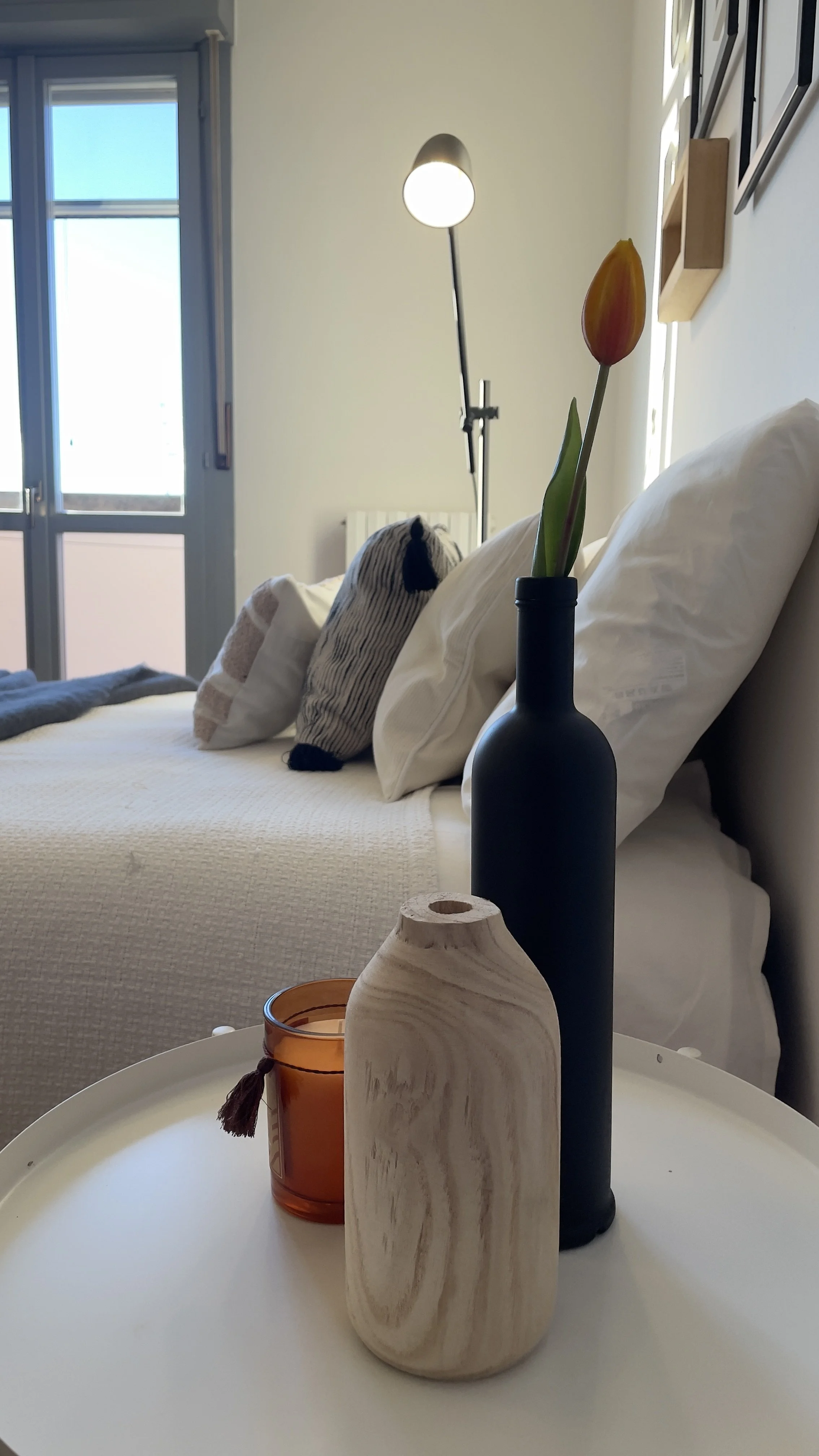 Interno di una camera da letto moderna con letto bianco, cuscini, lampada da lettura, bottiglia nera con tulipano arancione, bottiglia di legno e candela sul comodino.