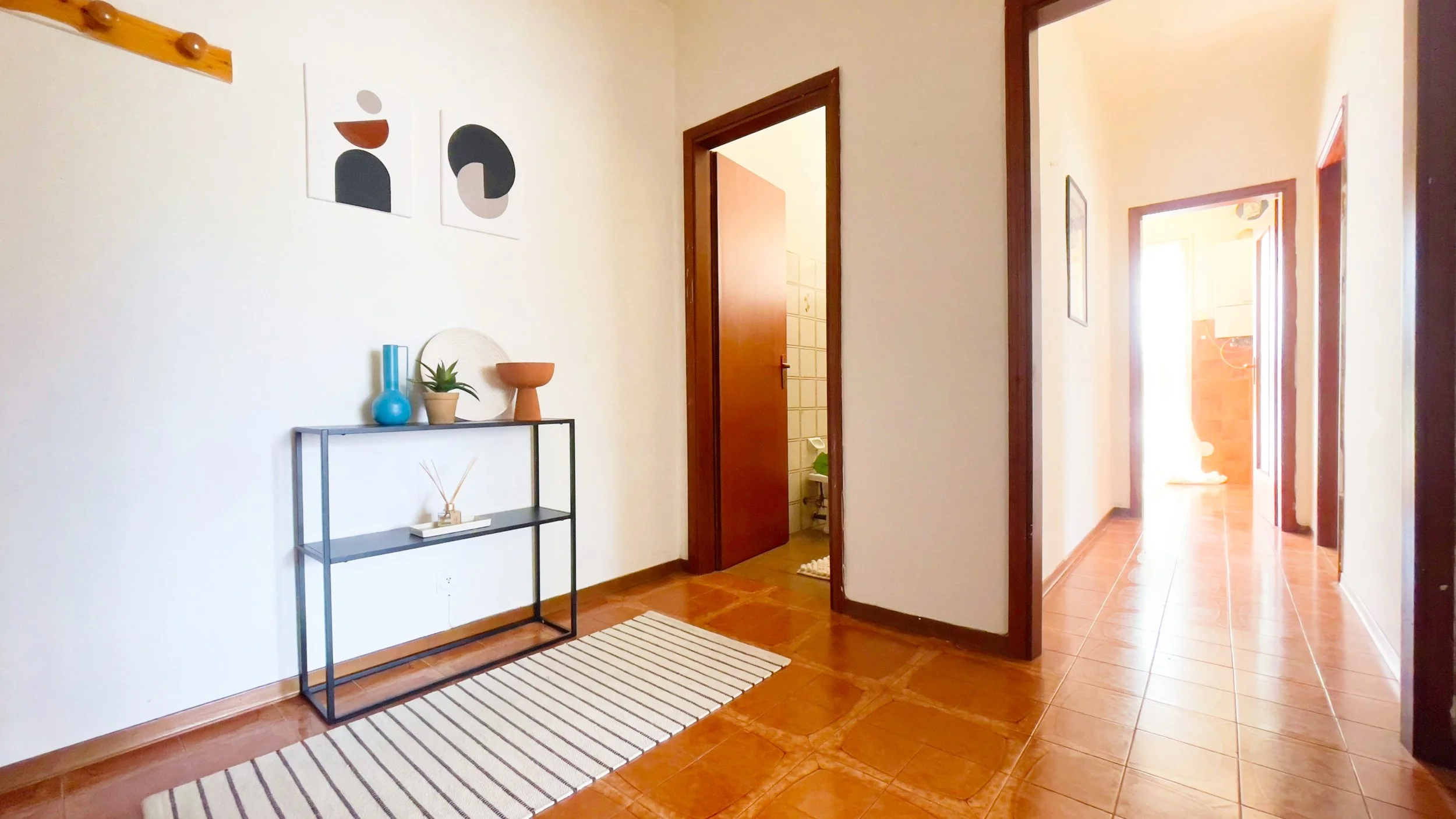 Corridoio di una casa con pavimento in piastrelle marroni, un tappeto a righe bianche e nere, un tavolo con oggetti decorativi come vasi e una pianta, e quadri astratti appesi sulla parete; porte aperte che conducono ad altre stanze.