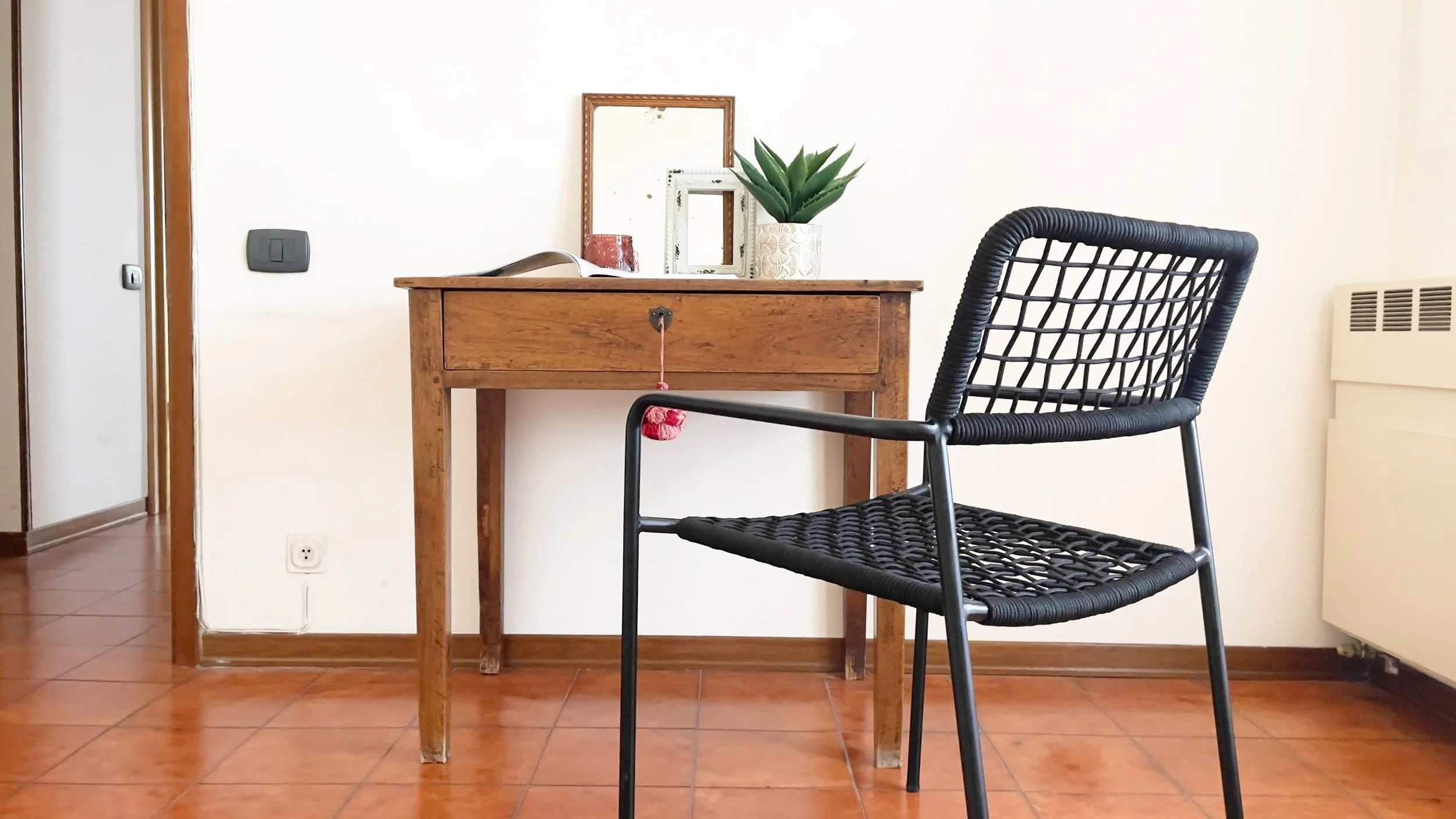Interno di una stanza con una sedia nera in primo piano e una scrivania in legno sullo sfondo. Sopra la scrivania ci sono una pianta in vaso, un libro aperto, e due cornici. Il pavimento è in piastrelle rosse.