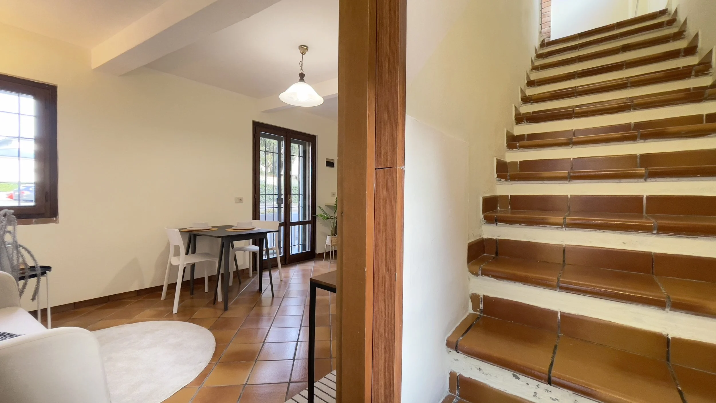 Interno di una casa con scale in legno, una zona pranzo con tavolo nero e sedie bianche, pavimento in piastrelle marroni e una finestra con persiane.