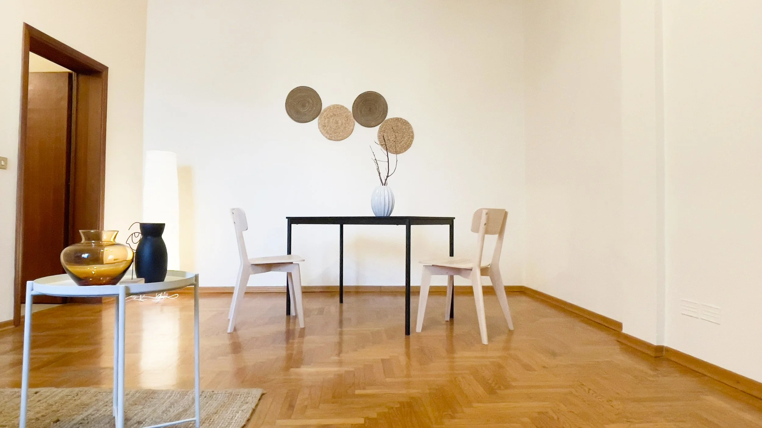 Stanza minimalista con pavimento in legno, due sedie beige, tavolo nero con vaso, e decorazioni a parete.