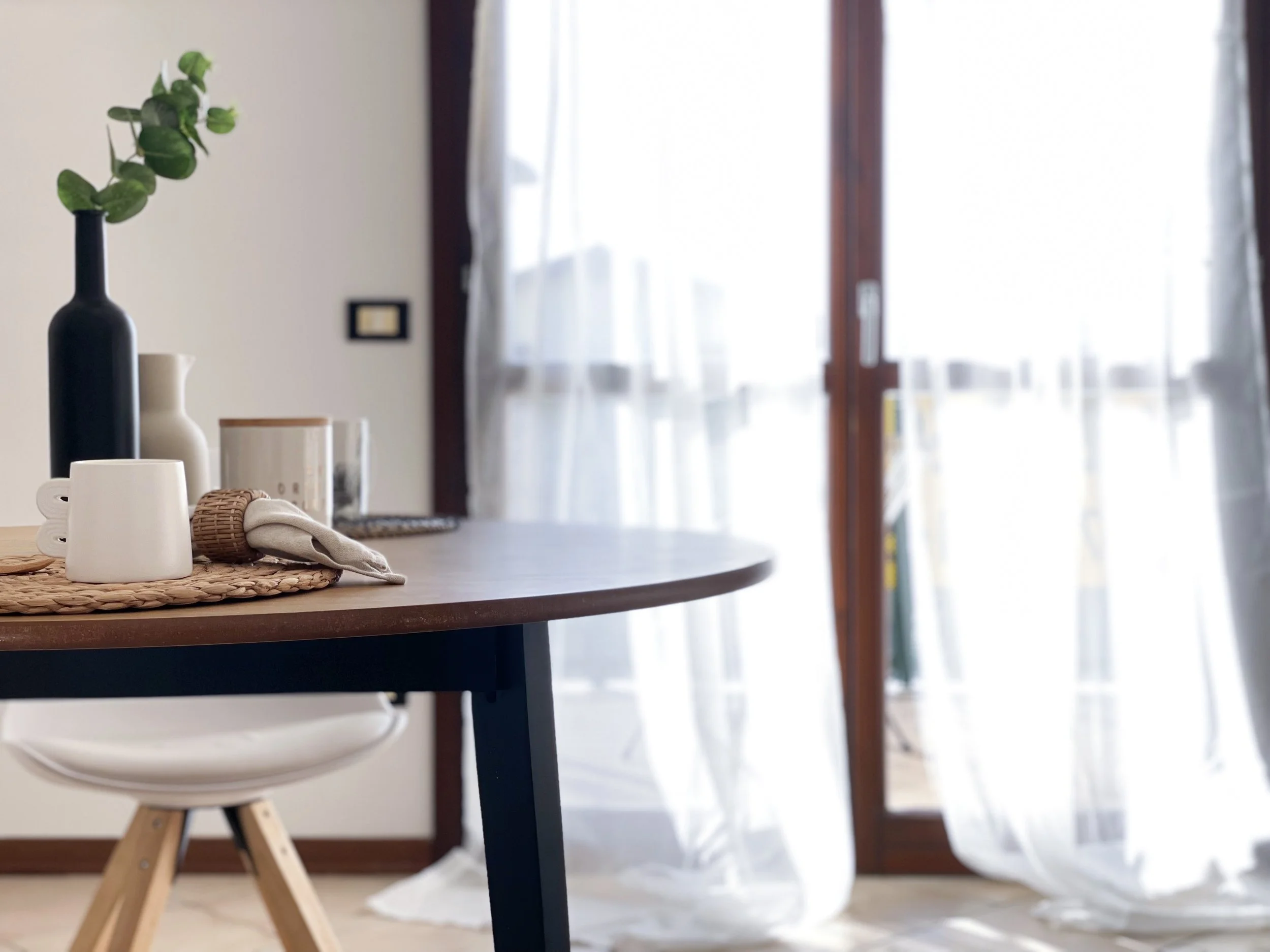 Interno di una stanza luminosa con una tavola rotonda in legno scuro, decorata con una caraffa bianca, un vaso nero con rami verdi e una tazza bianca. Sullo sfondo, una finestra con tende bianche trasparenti. Una sedia con gambe in legno è visibile s