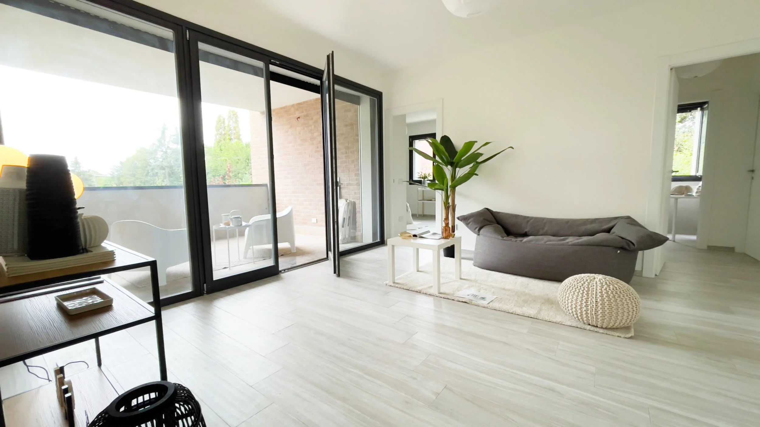 Interno di un soggiorno moderno con pavimento di legno chiaro, divano grigio, tavolino bianco con pianta, e grande finestra che dà su un balcone con sedie bianche.