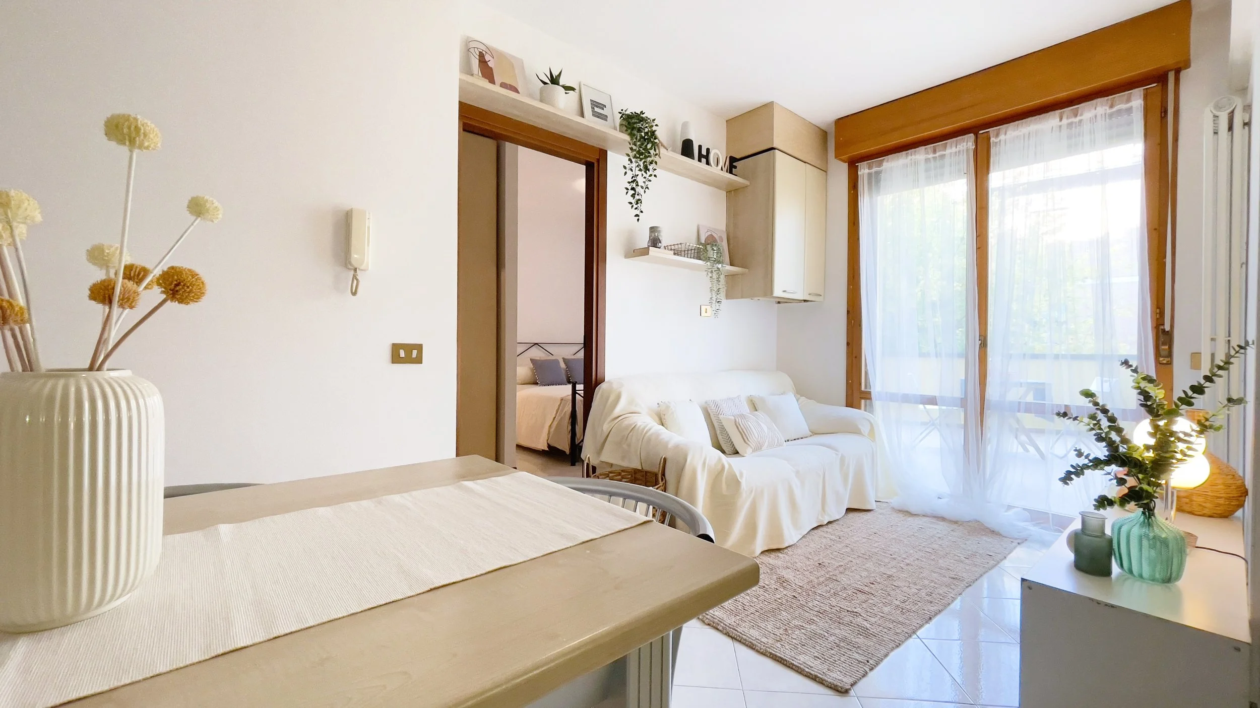 Interno di un soggiorno luminoso con divano bianco, tavolino con decorazioni floreali, porta finestra con tende bianche, e accessori decorativi sugli scaffali.