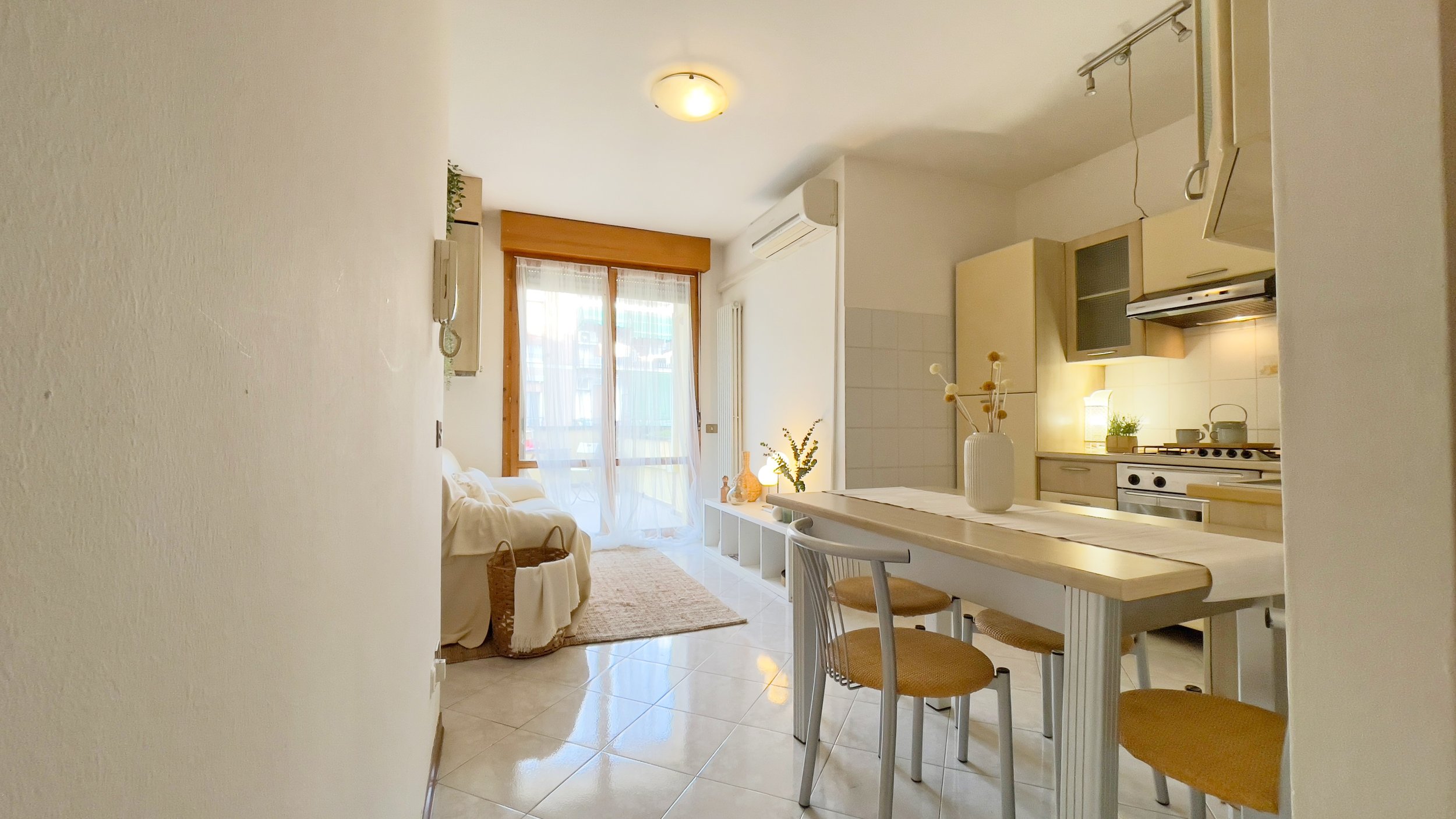 Interno di un appartamento moderno con cucina a vista, tavolo da pranzo e soggiorno con divano e finestre ampie.