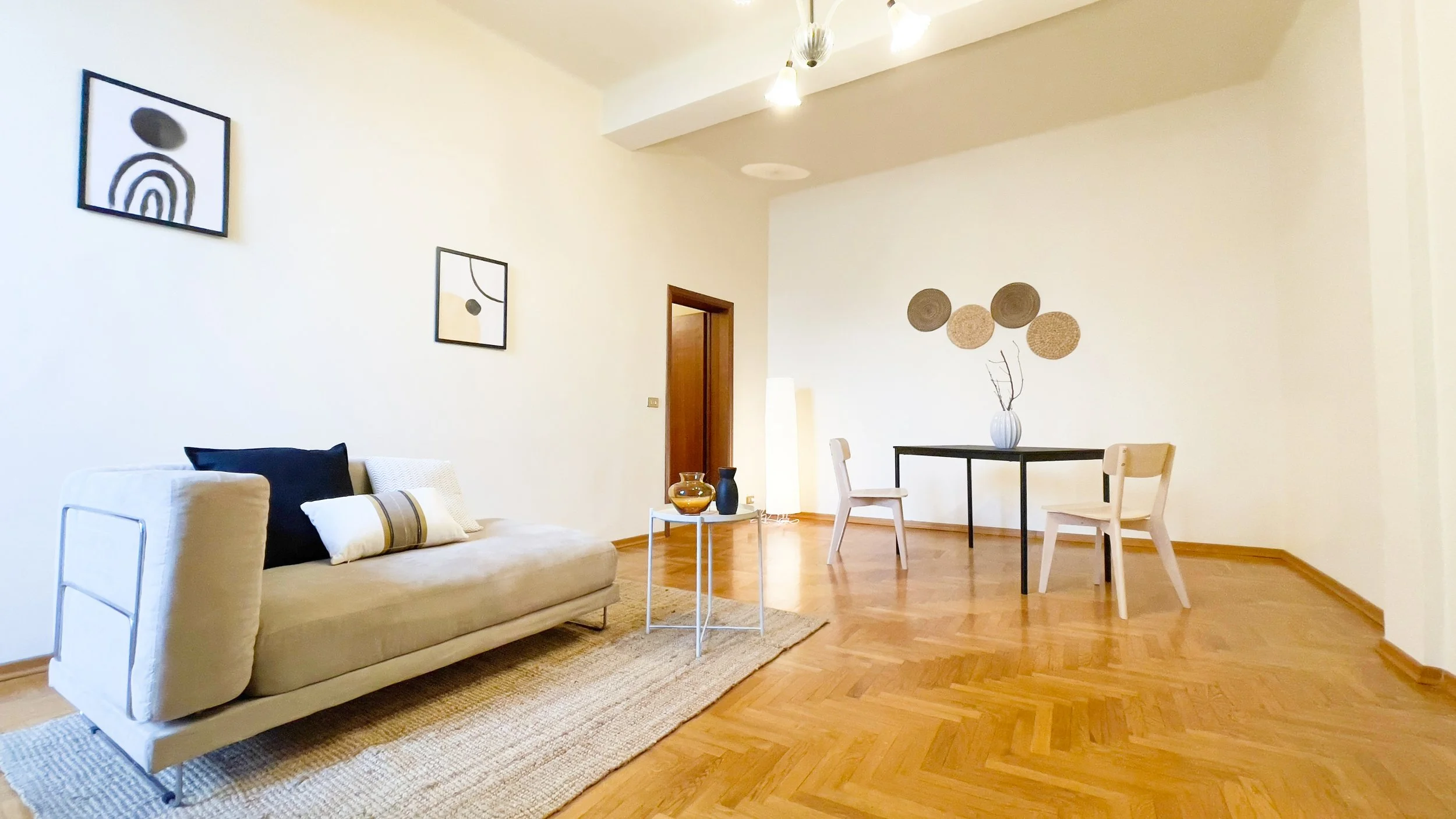 Interno di un soggiorno moderno con divano beige, tavolino bianco, tavolo nero con sedie in legno chiaro, cornici astratte alle pareti e decorazioni circolari. Pavimento in legno chiaro.