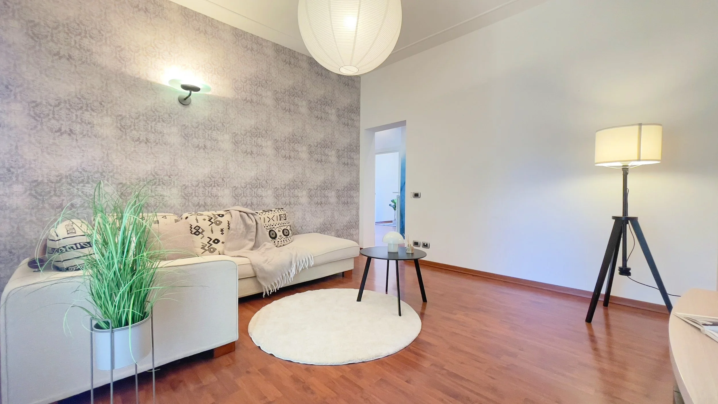 Soggiorno moderno con divano bianco, tavolino nero, pianta decorativa, lampada da terra e carta da parati geometrica grigia.