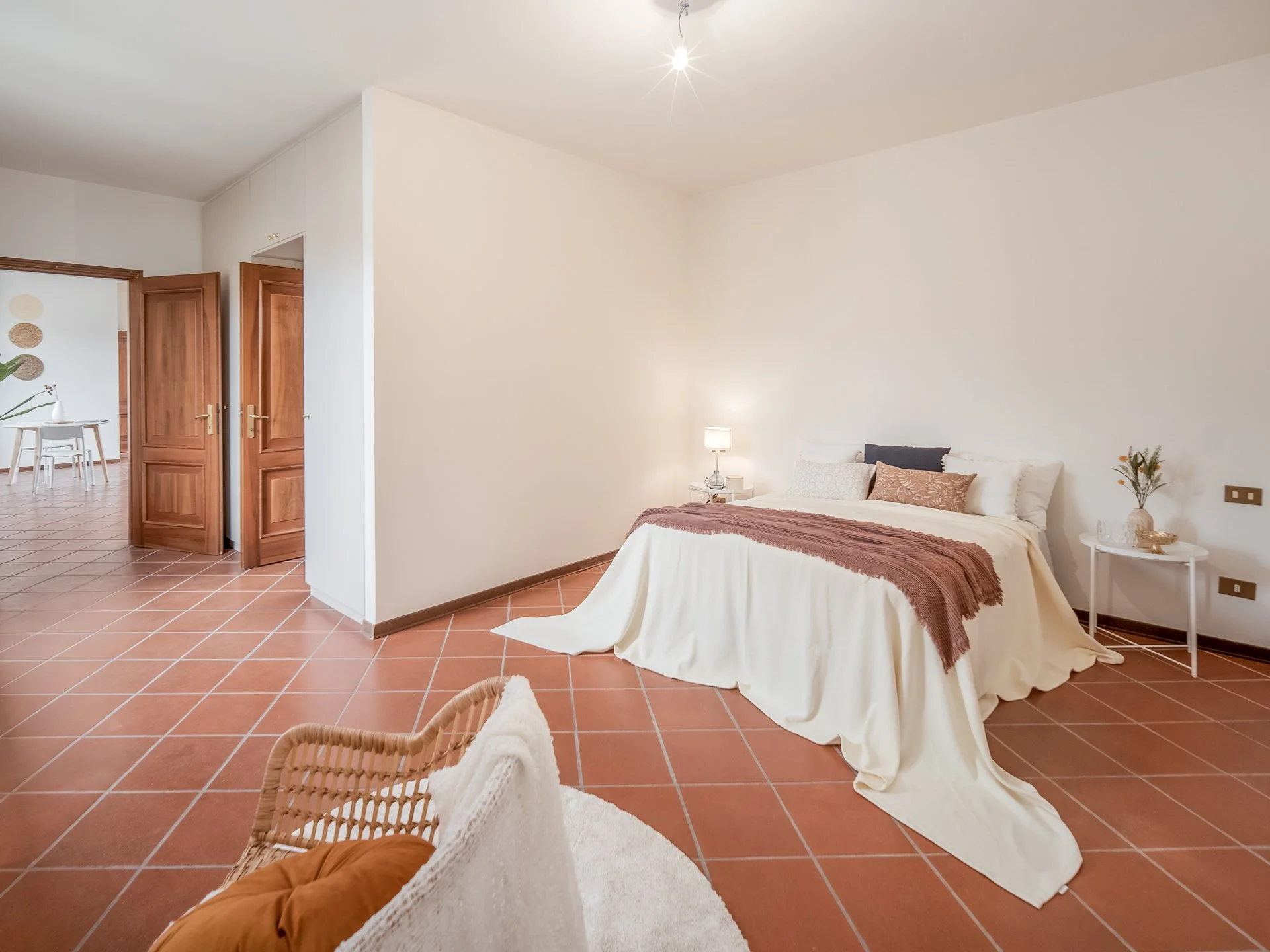 Camera da letto con pavimento in cotto, letto matrimoniale con copriletto bianco e cuscini, tavolino rotondo con lampada e decorazioni, ambiente luminoso con porte in legno aperte verso un'altra stanza.