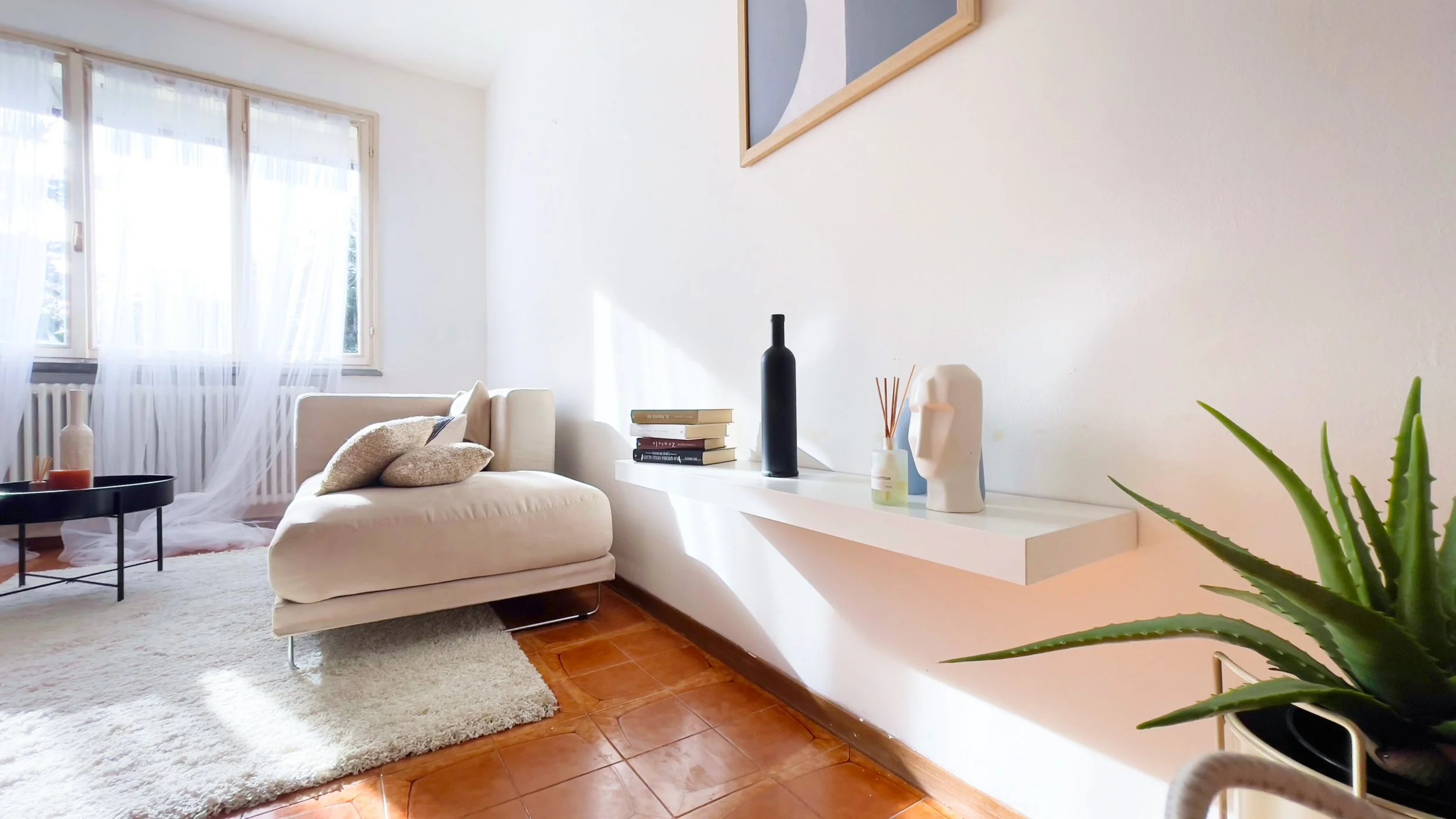 Soggiorno moderno con divano beige, tavolino nero circolare, scaffale bianco con decorazioni, finestra con tende trasparenti, pianta in vaso e pavimento in piastrelle marroni.