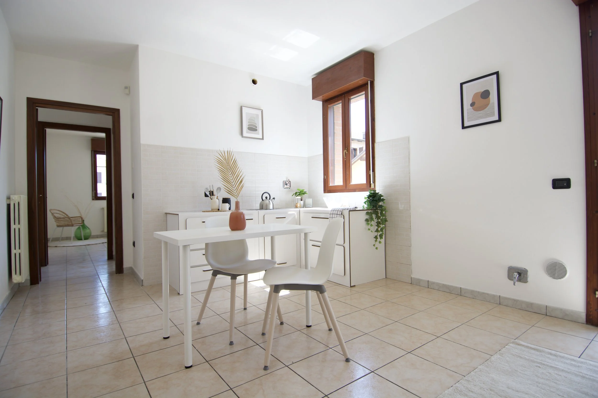 Cucina moderna con pareti bianche, tavolo e sedie bianche, decorazioni minimaliste, pianta verde su un mobile, e finestra aperta con cornice marrone.