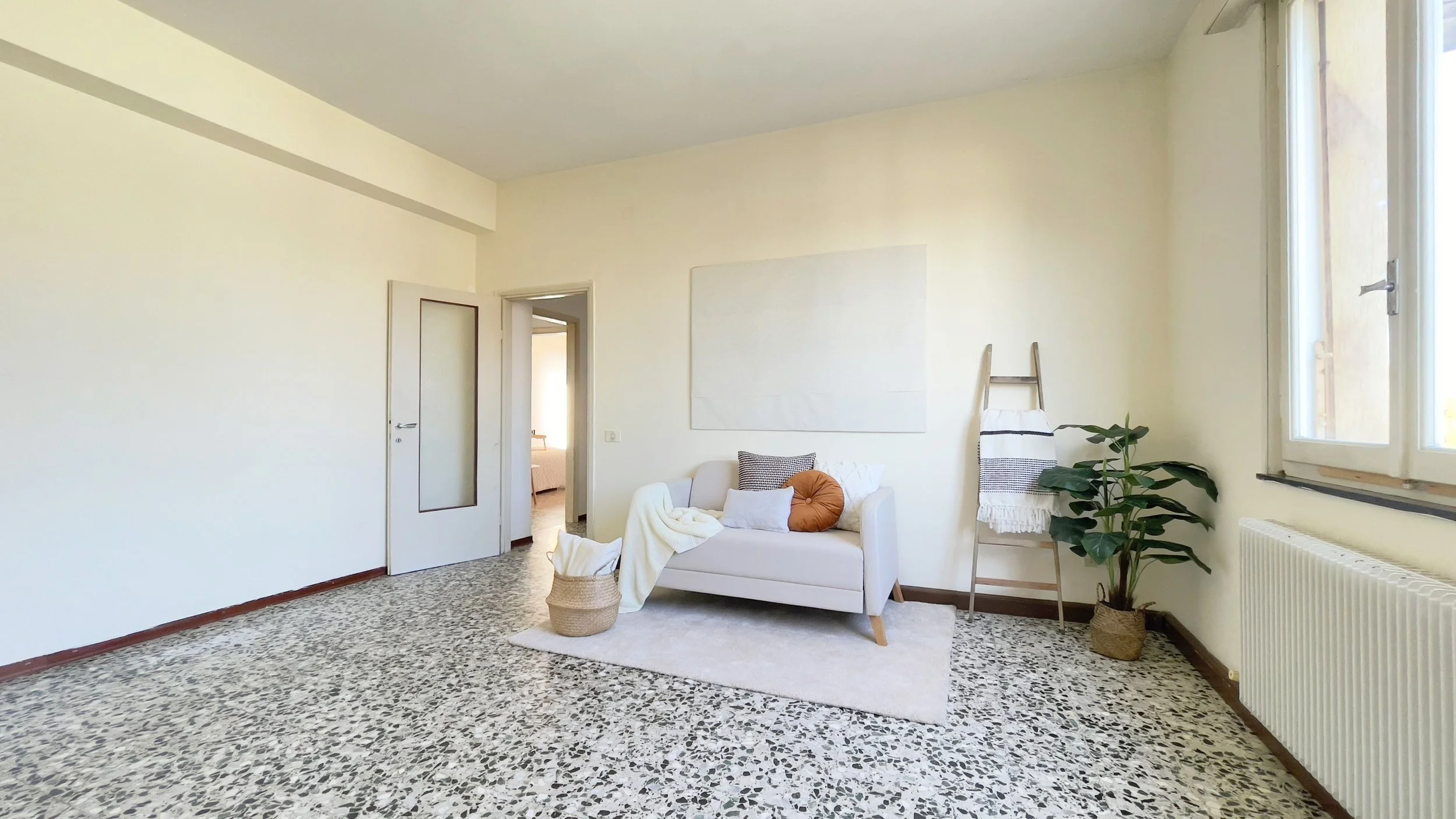 Soggiorno luminoso con divano bianco, cuscini, pianta decorativa e scaletta porta asciugamani.