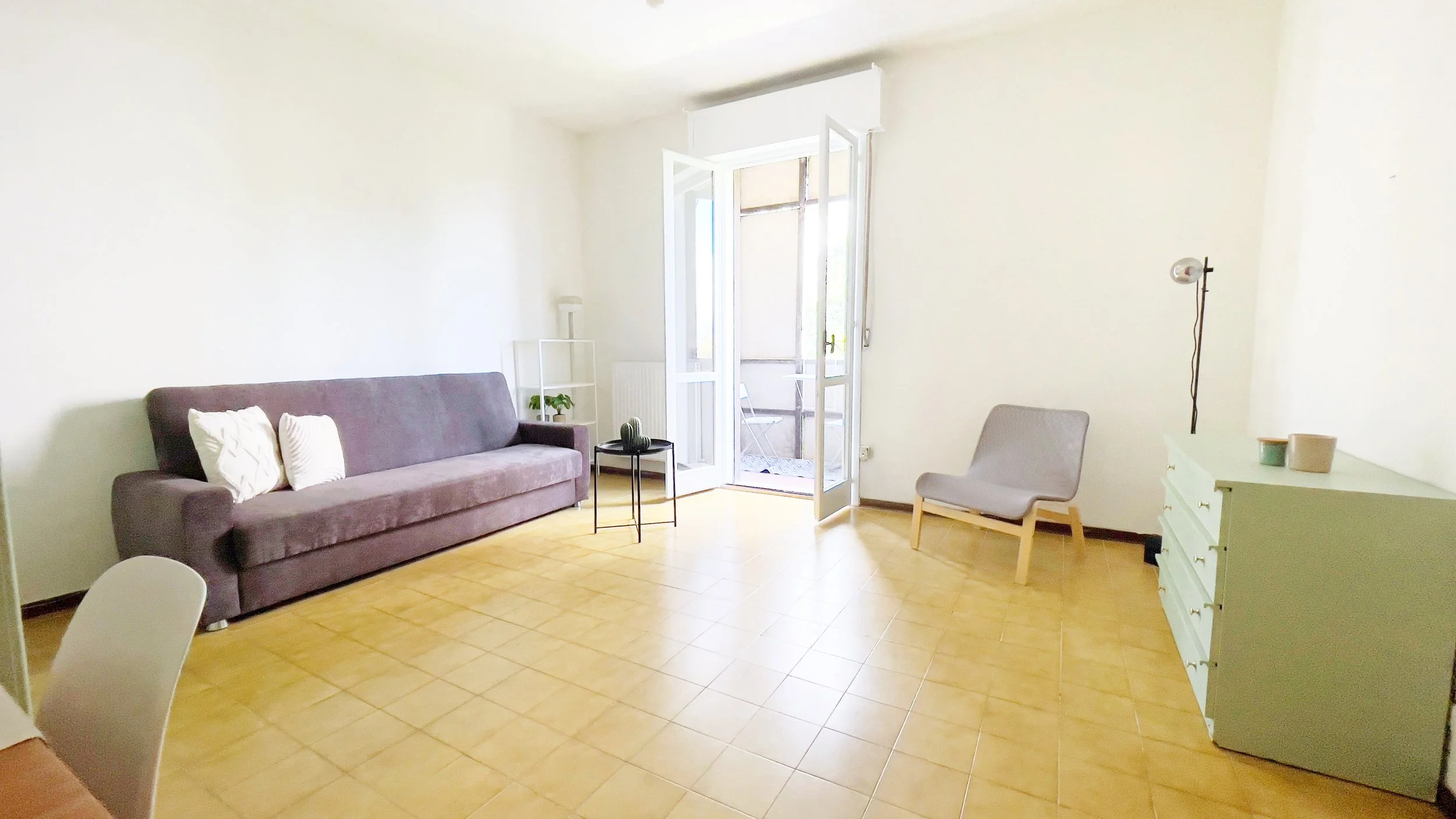Soggiorno luminoso con divano grigio, sedia moderna, cassettiera verde, tavolino nero, porta-finestra aperta e pavimento in piastrelle gialle.