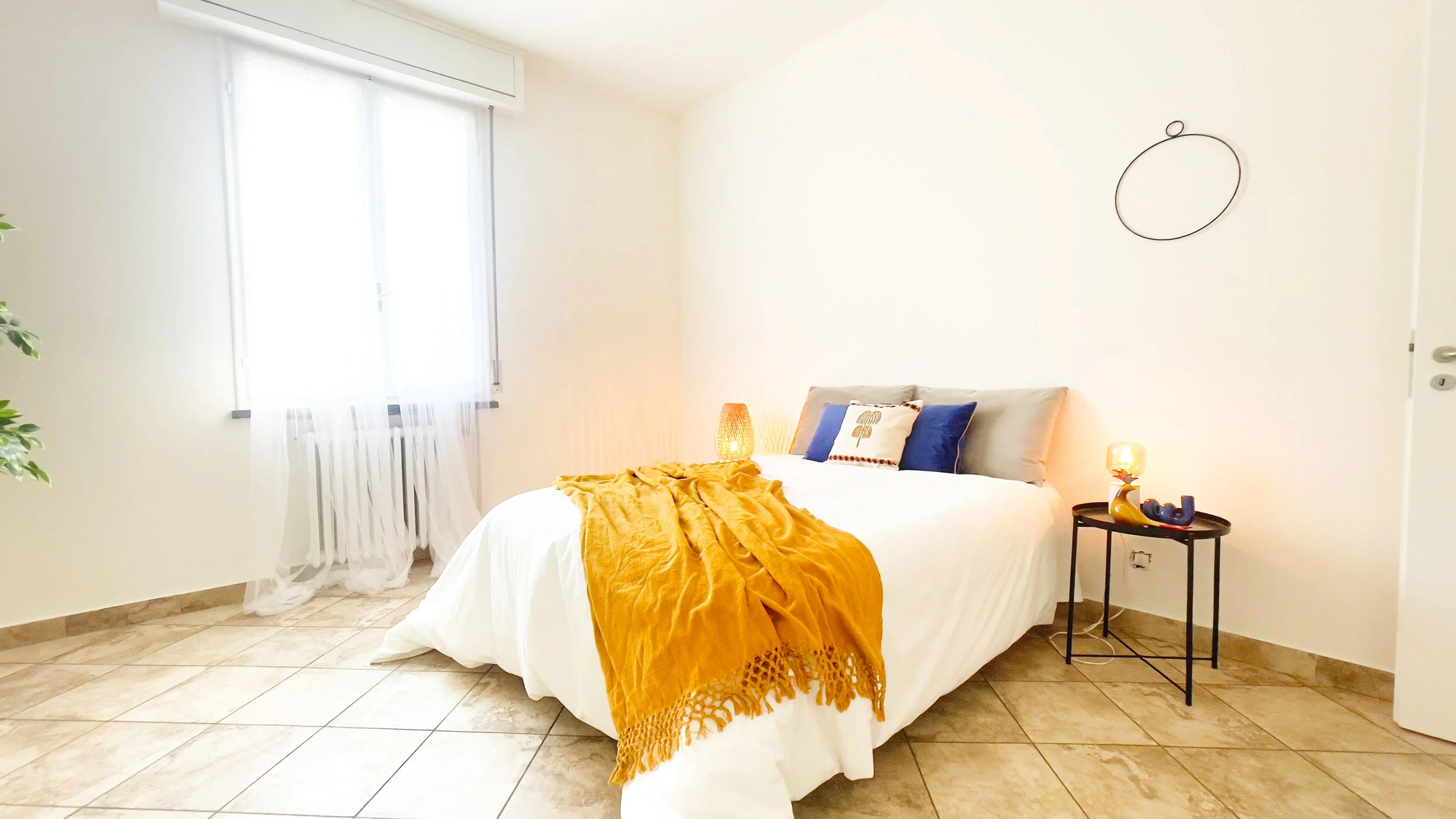 Camera da letto minimalista con letto matrimoniale, coperta arancione, cuscini decorativi e lampada su comodino.