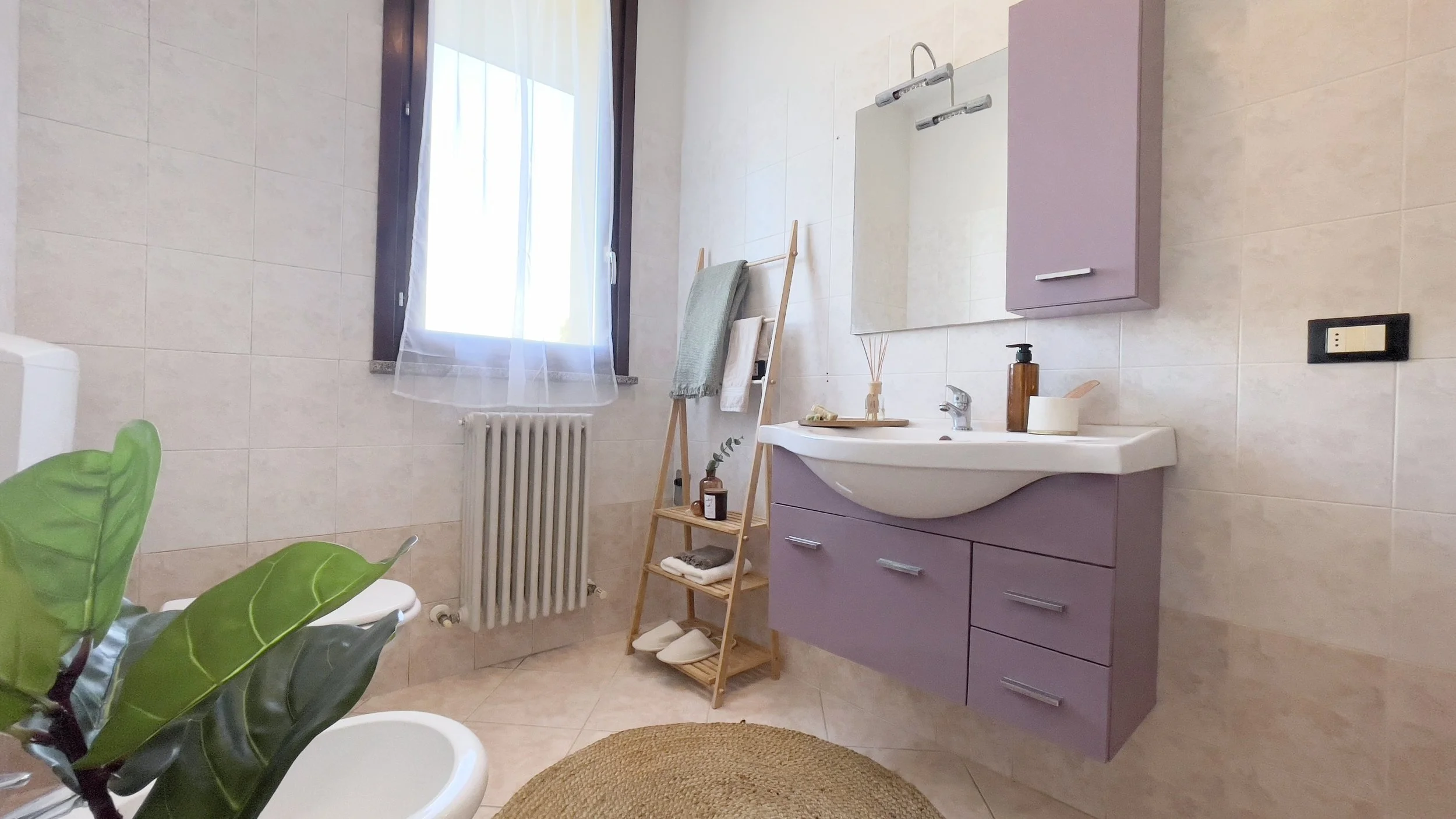 Bagno moderno con lavandino e mobiletti viola, scaffale in legno con asciugamani, finestra con tende, e pianta decorativa. Pavimento piastrellato chiaro e tappeto rotondo di paglia.